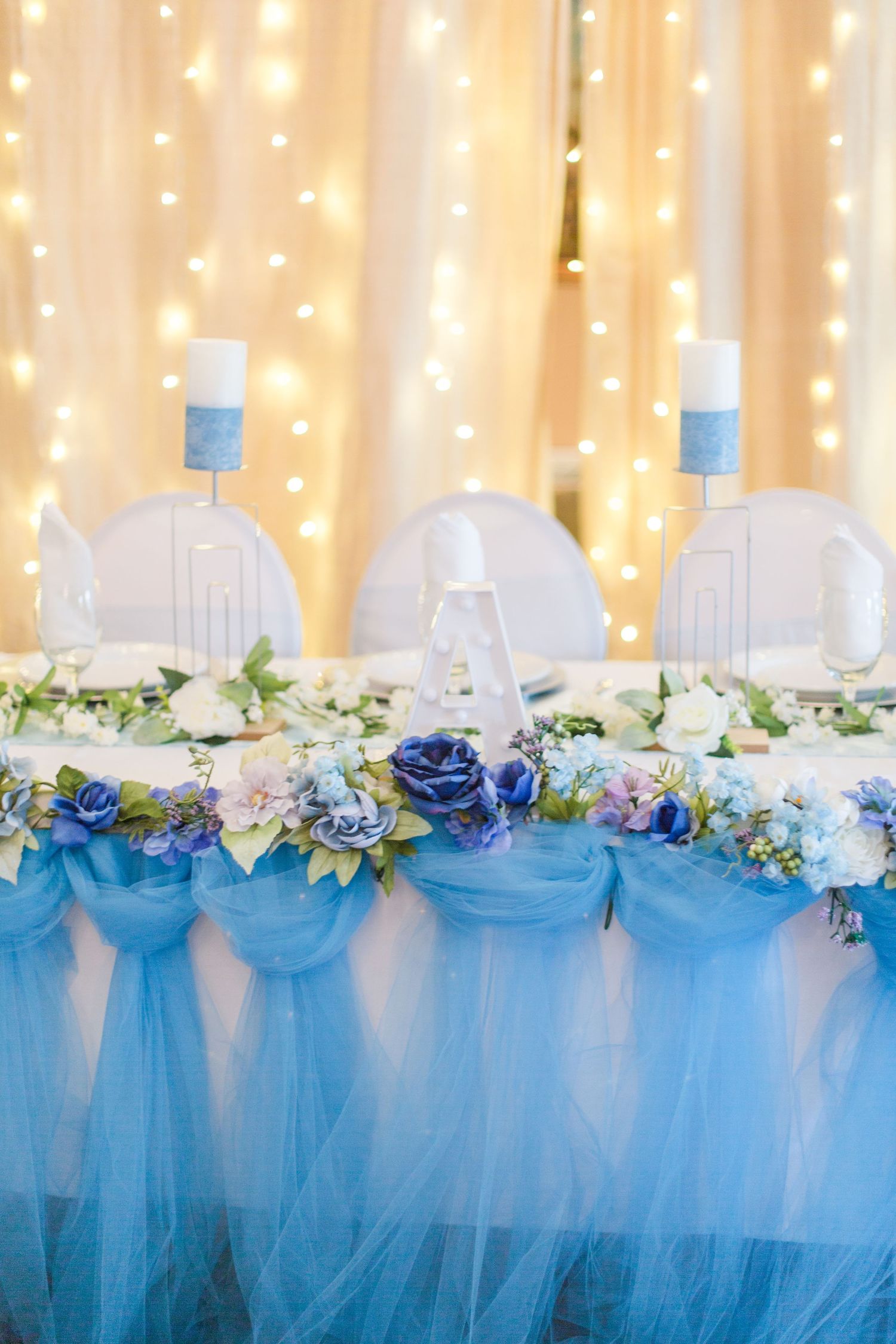 Wedding reception head table decorated with blue tulle skirting, white flowers and twinkling fairy lights.