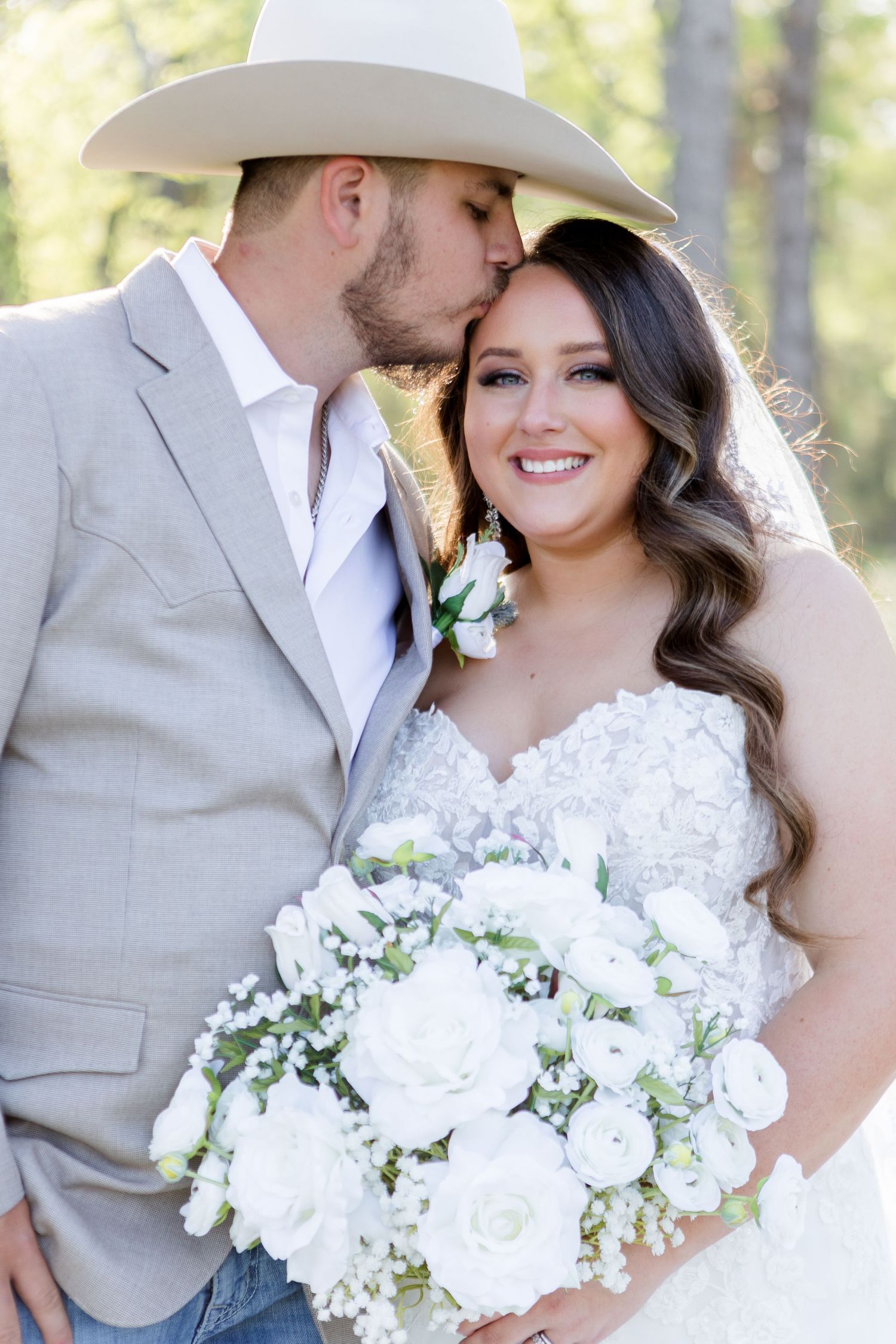 Wedding couple in rustic attire with white floral bouquet posing among trees.