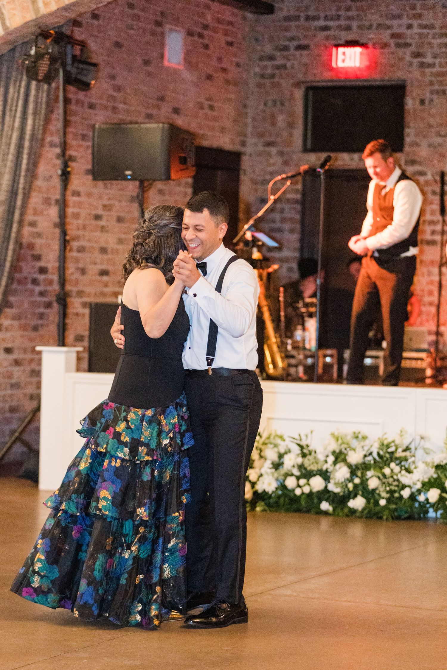 Wedding dance scene with colorful floral dresses against rustic brick backdrop.