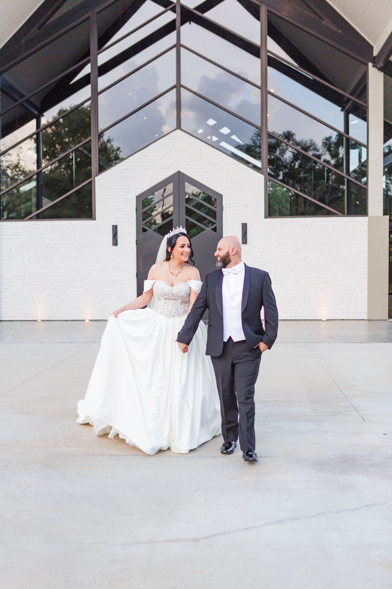A couple in formal wedding attire take posed photos against a modern white building exterior.
