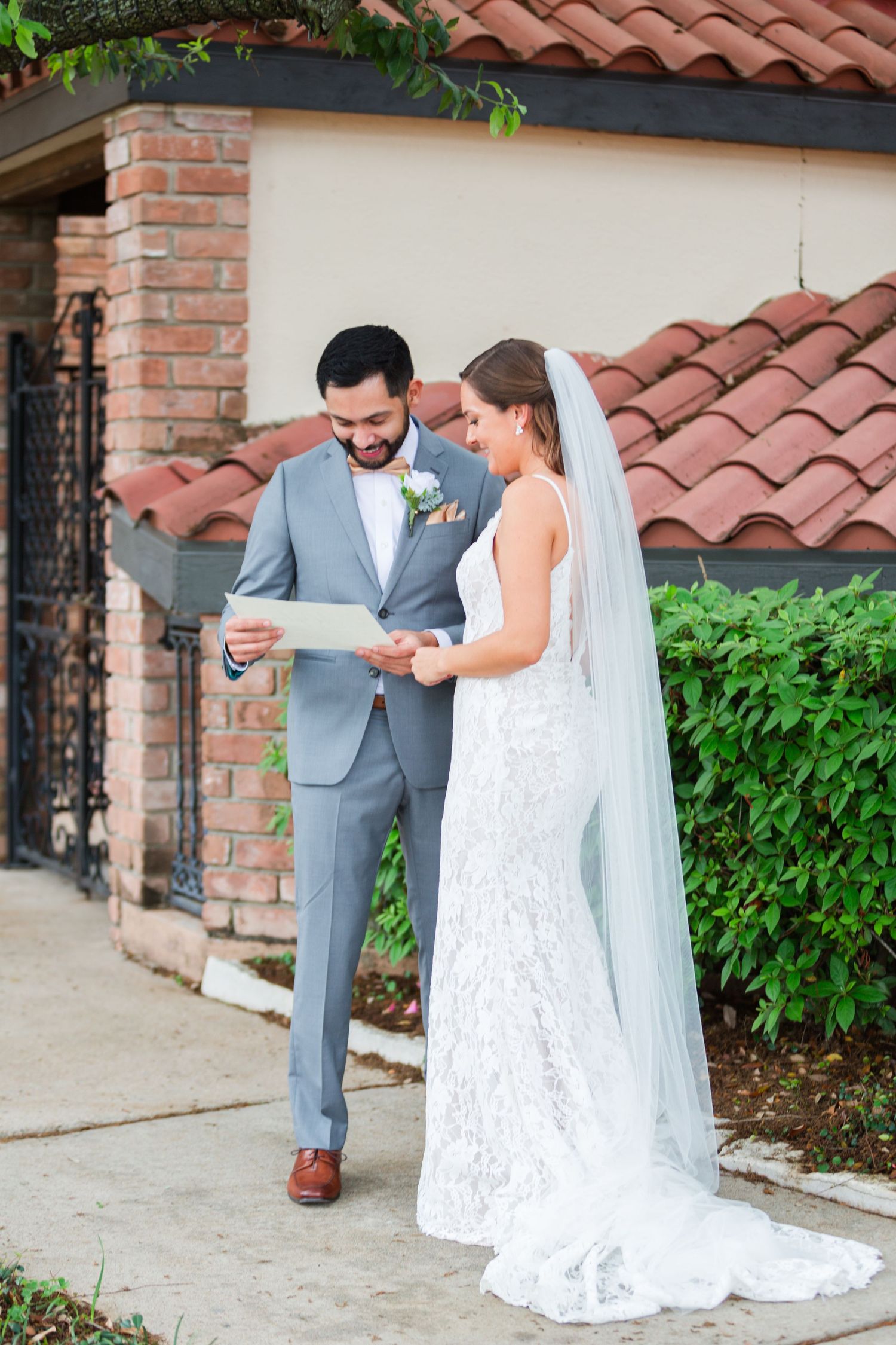 Wedding couple in grey suit and lace dress sharing first look moment by brick building.