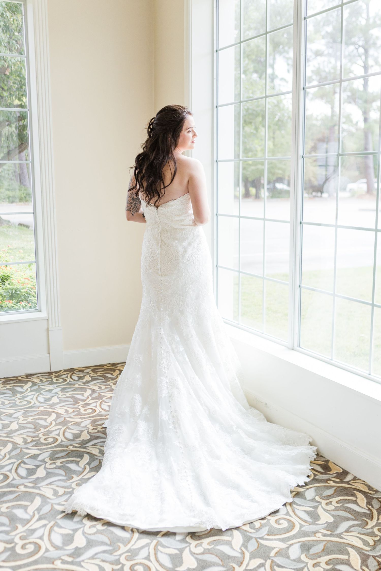 A bride in a white lace wedding dress poses gracefully by large windows with an ornate patterned floor below.