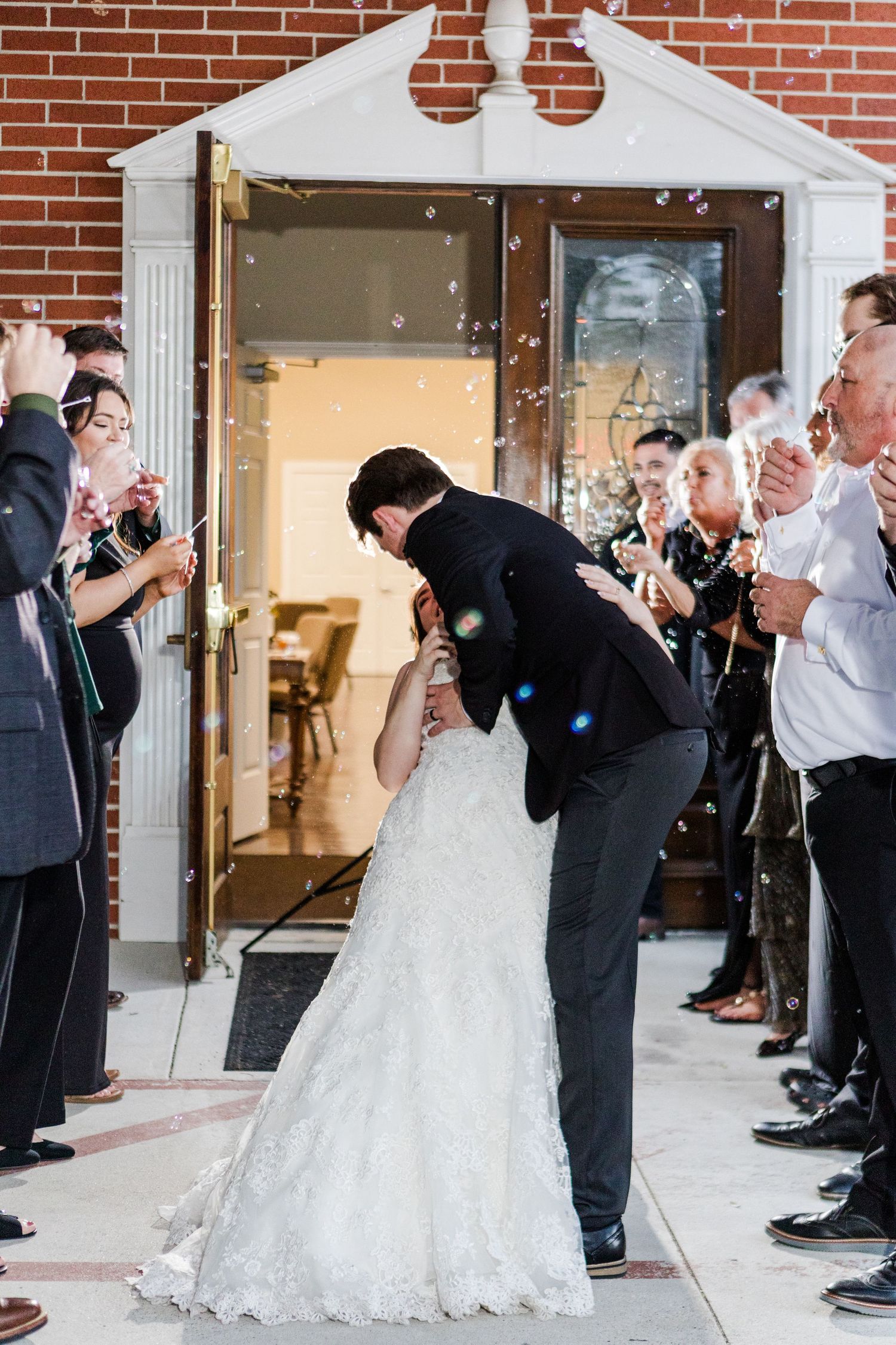 A newlywed couple shares their first kiss while guests throw confetti outside a brick church entrance.