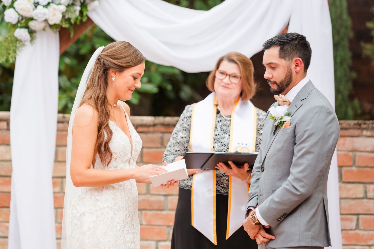 Wedding ceremony participants stand in front of a decorated brick wall with white draping and flowers.