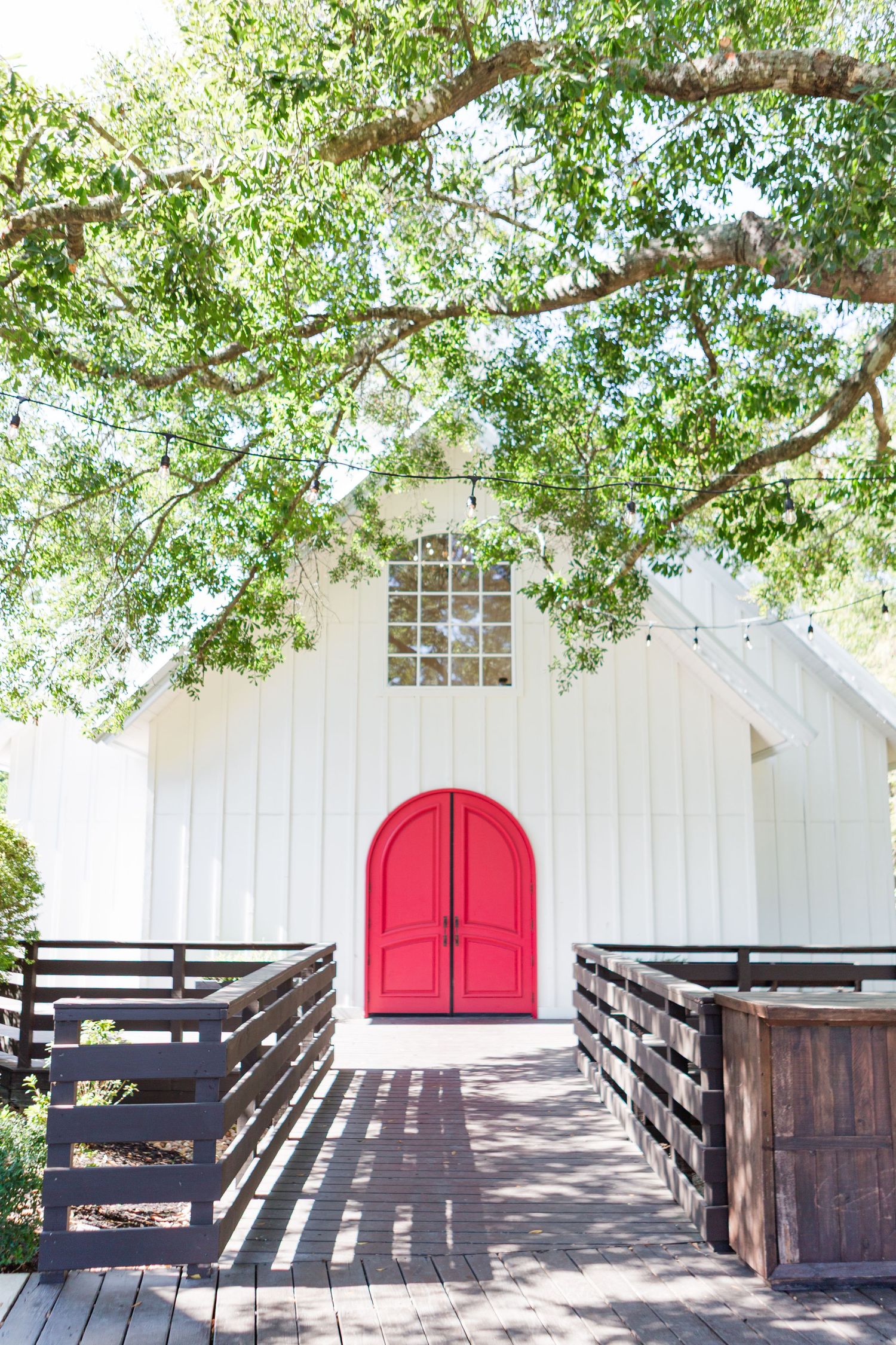 White chapel building with vibrant red double doors and wooden walkway under green tree branches.