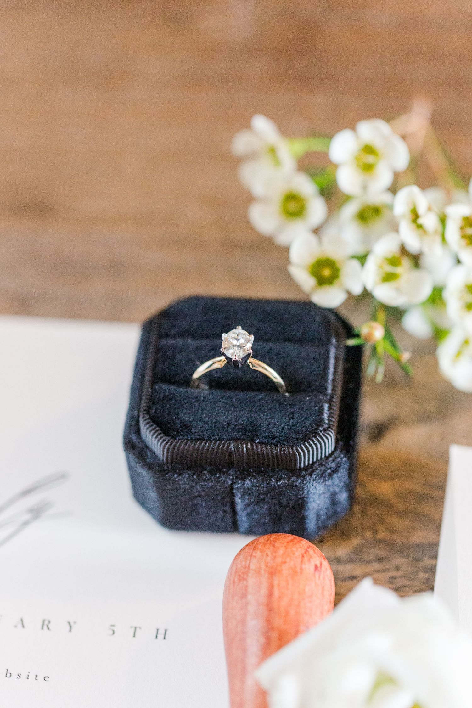 Diamond engagement ring displayed on black velvet ring box with delicate white flowers in background.