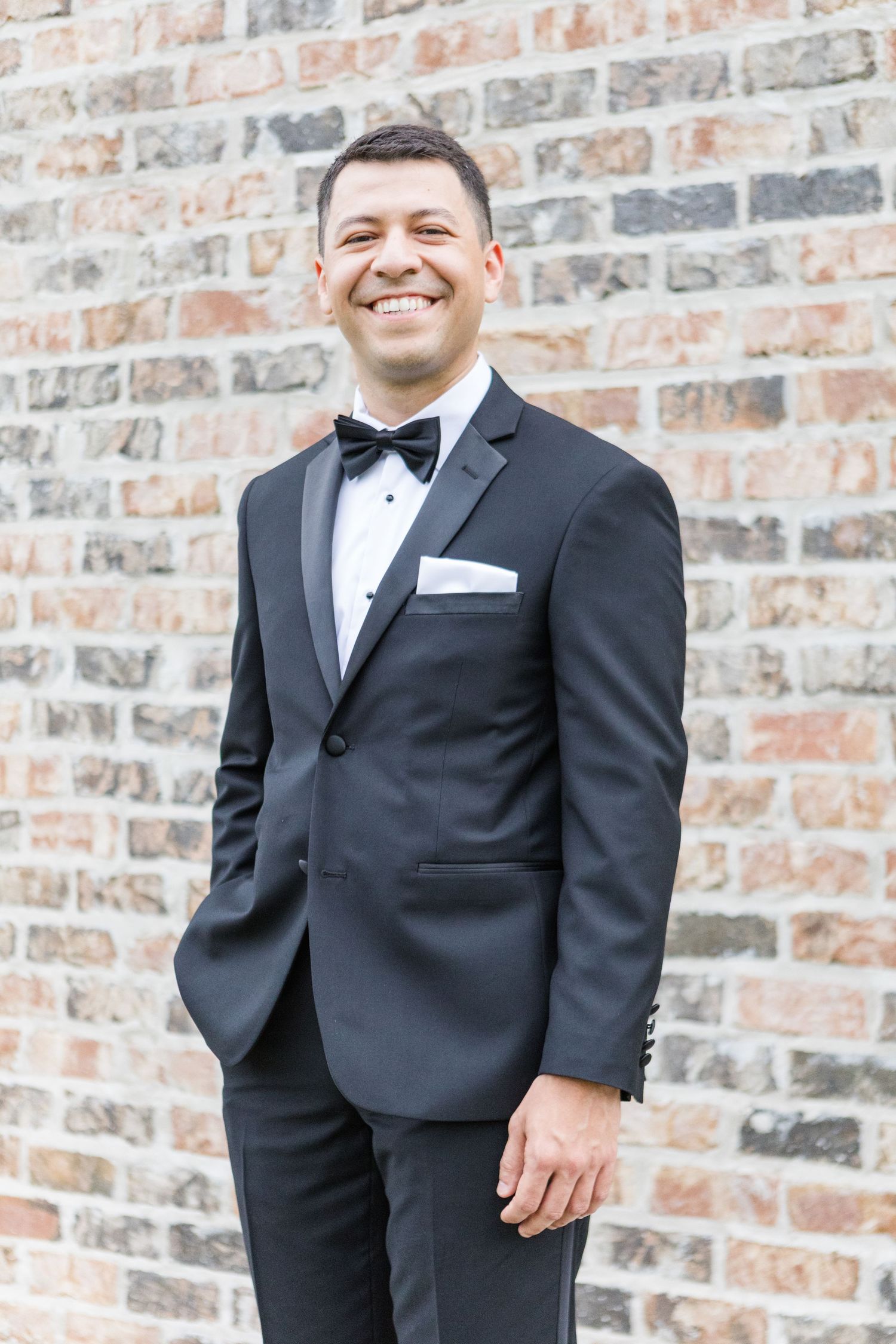 A groom in a classic black tuxedo and bowtie smiles against a rustic brick wall on his wedding day.