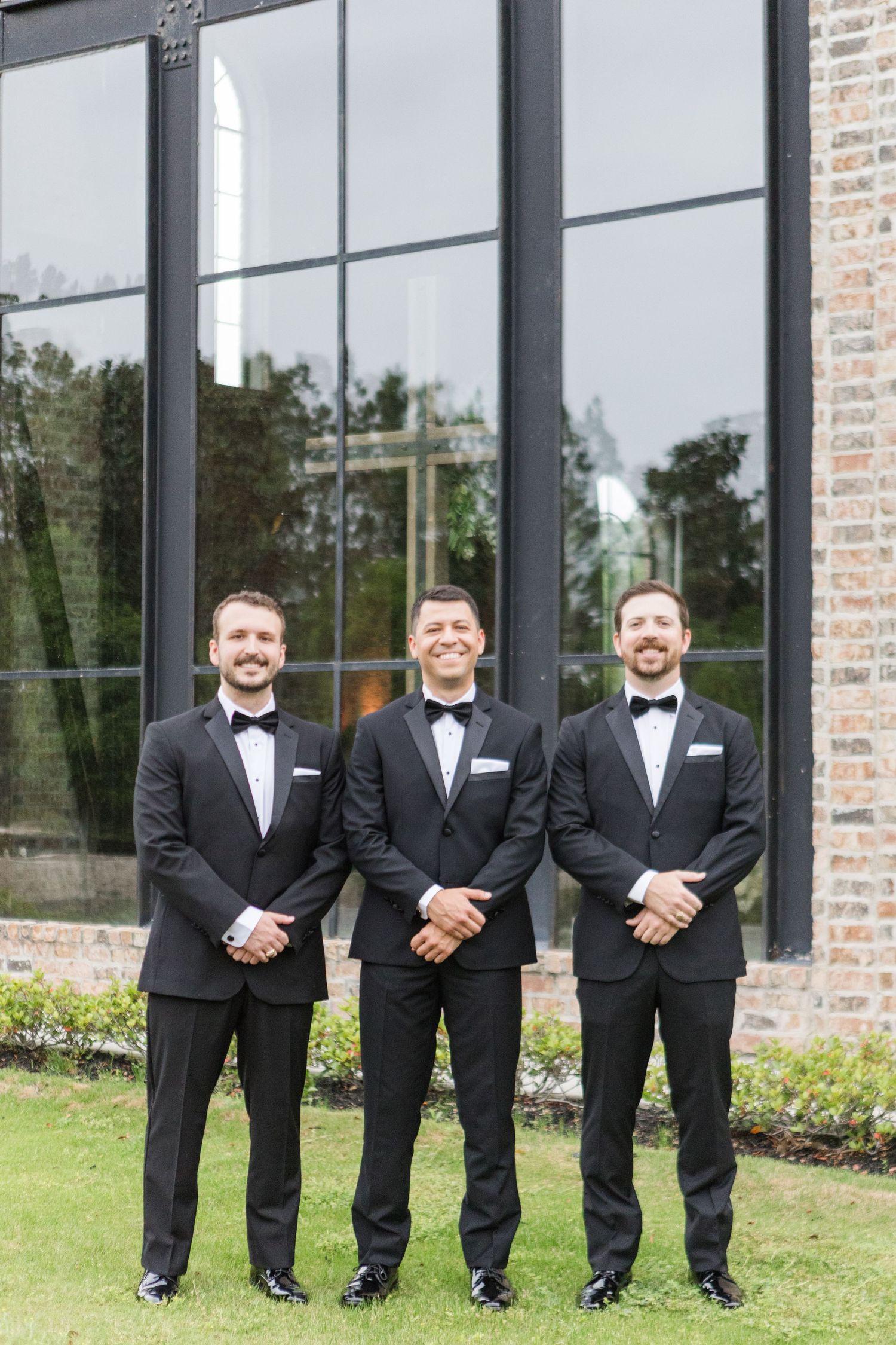 Groomsmen in matching tuxedos pose in line against brick building exterior.