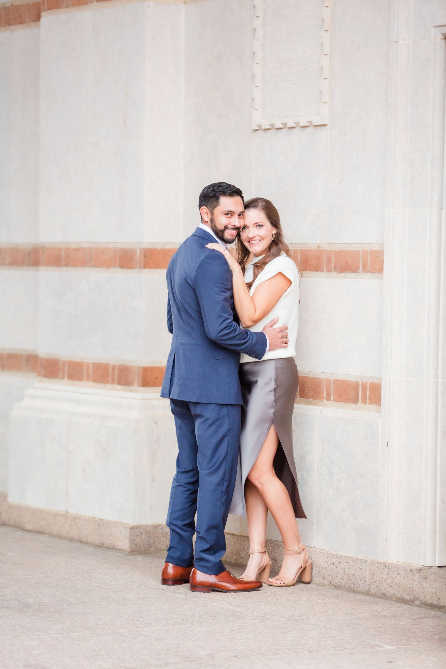 A couple in formal attire embraces against a light colored wall with decorative trim.