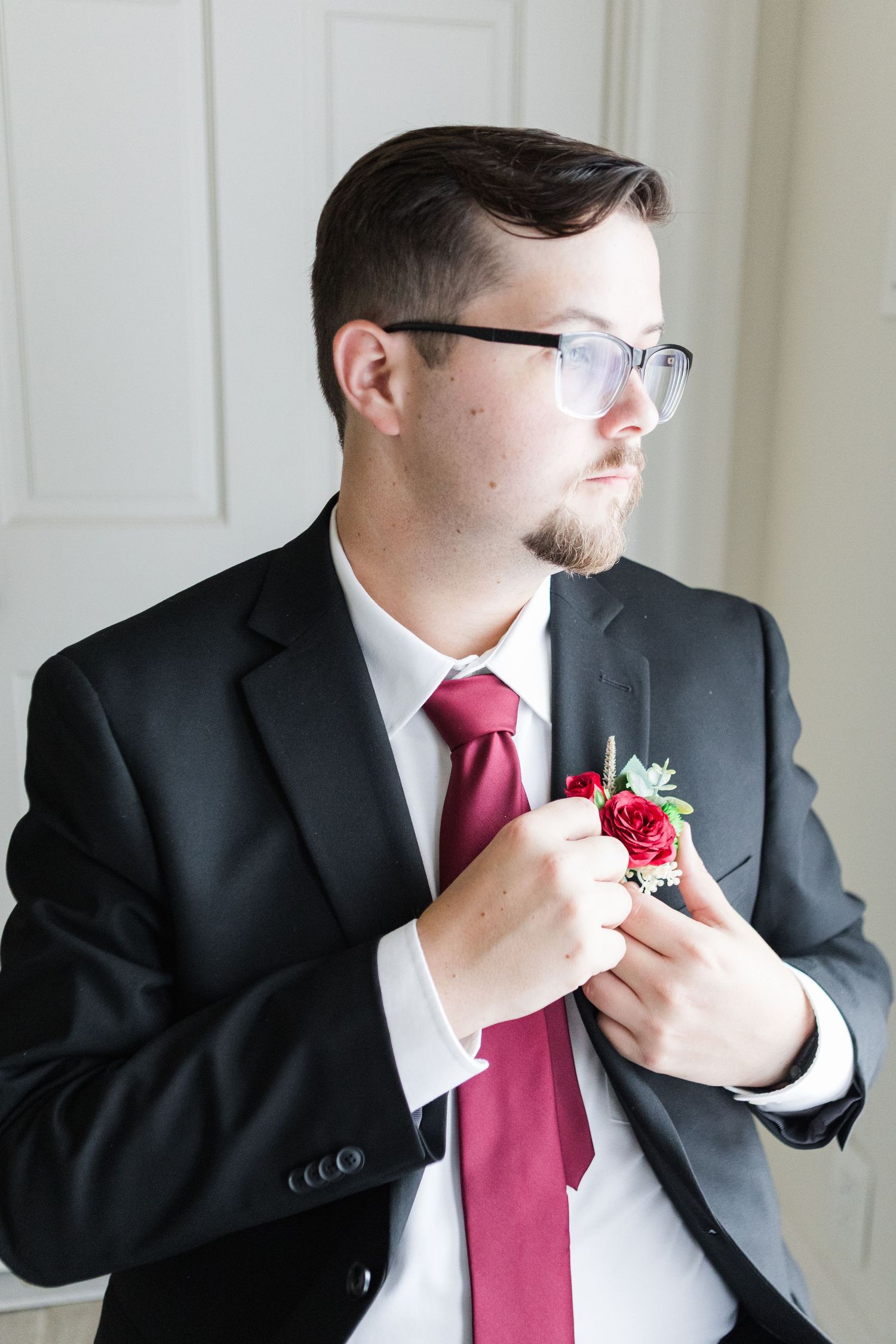 Groom in black suit and red tie adjusting boutonniere against white wall.