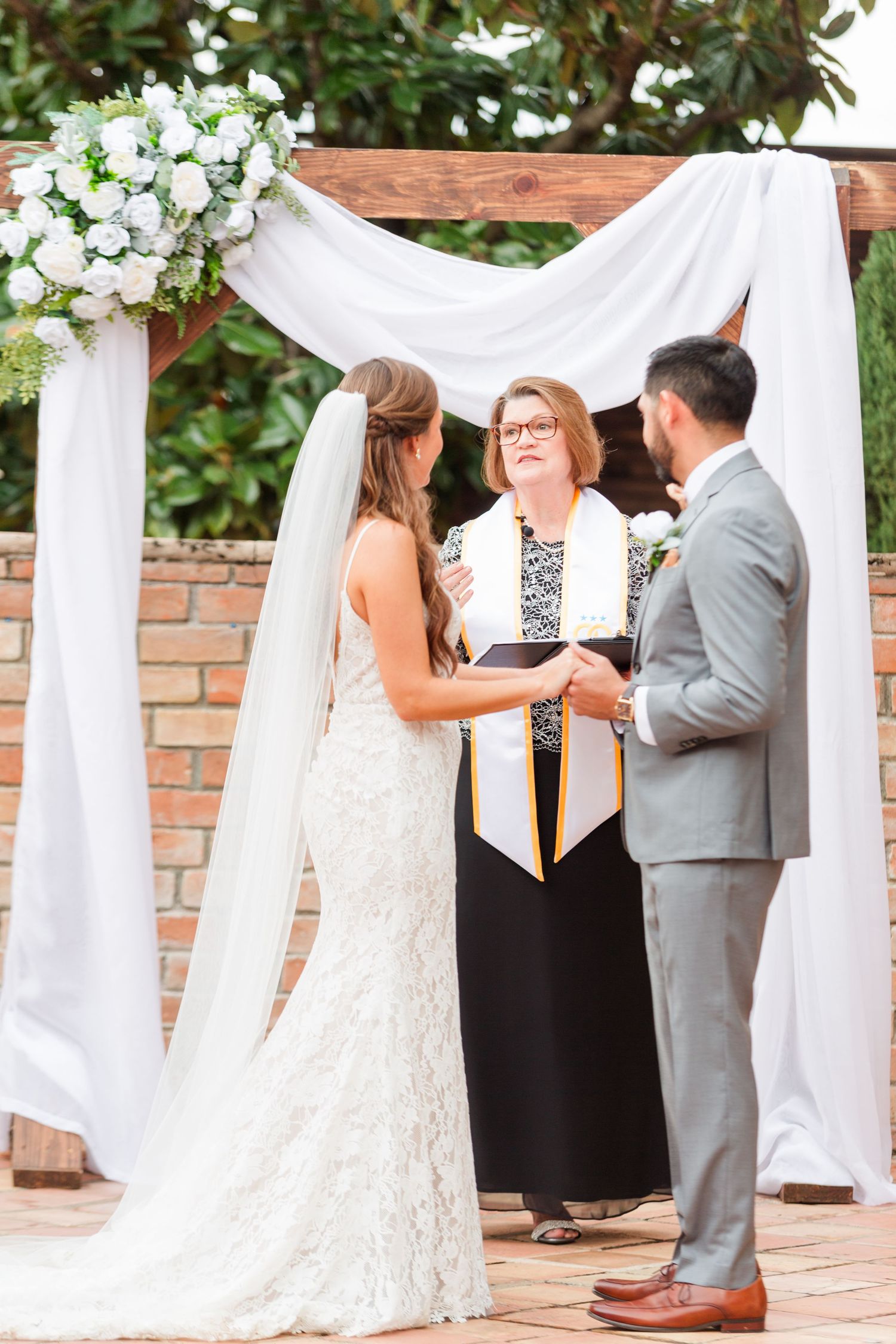 A wedding ceremony takes place under an arch decorated with white fabric and floral arrangements.