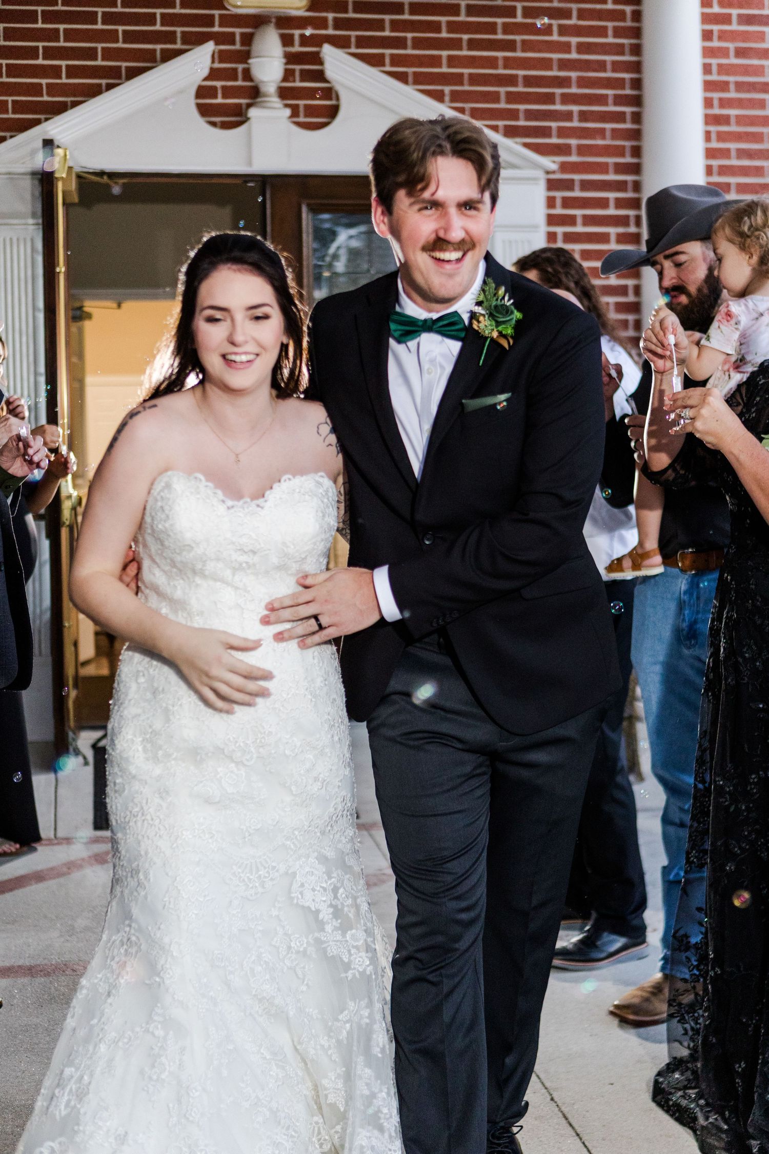 A newlywed couple celebrates joyfully outside a brick building with white columns.