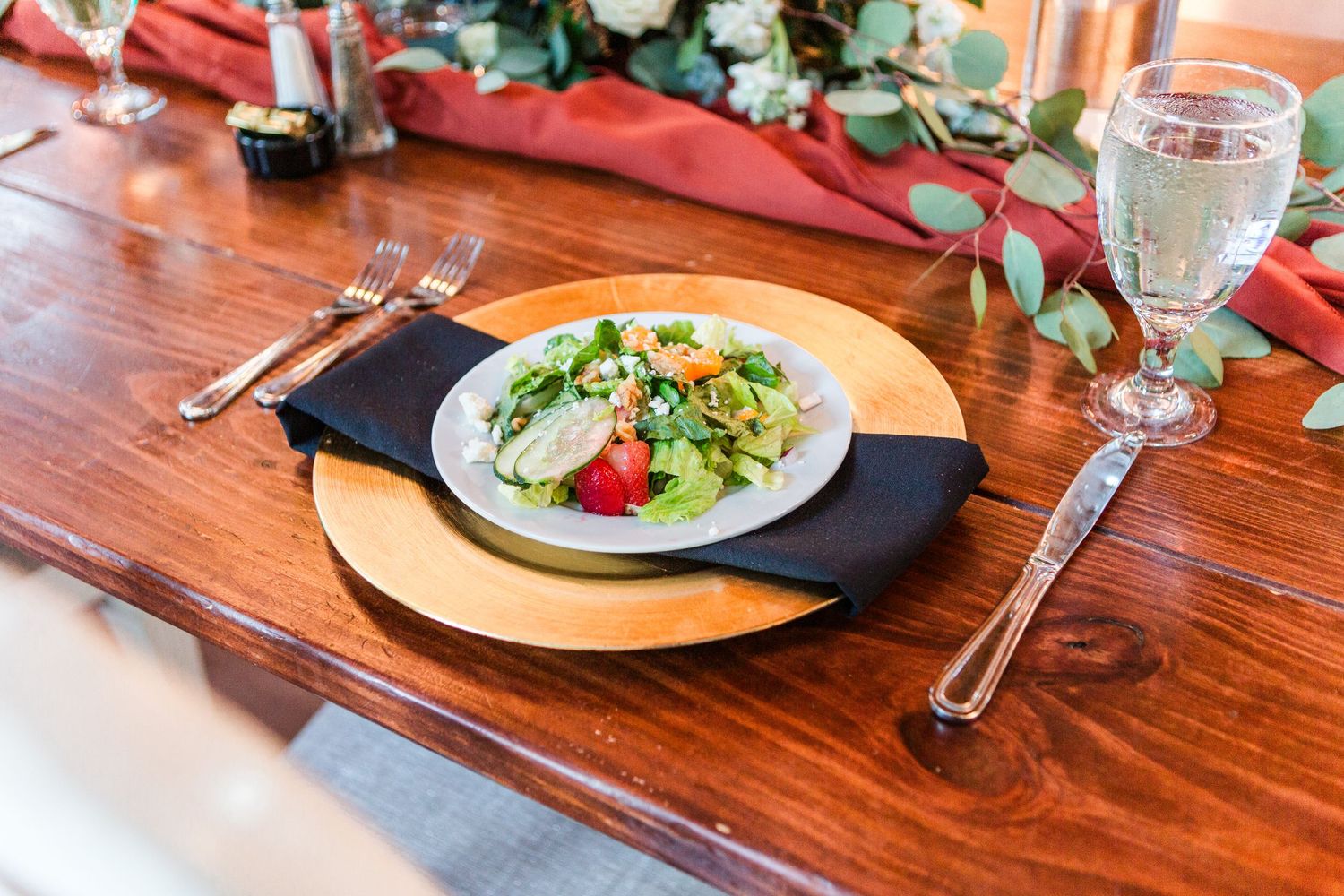 Close-up of wedding reception place settings with red napkins and greenery on wooden tables.