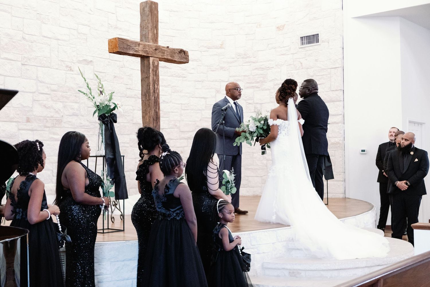 A wedding ceremony takes place in a white-walled church space with guests watching the couple at the altar.