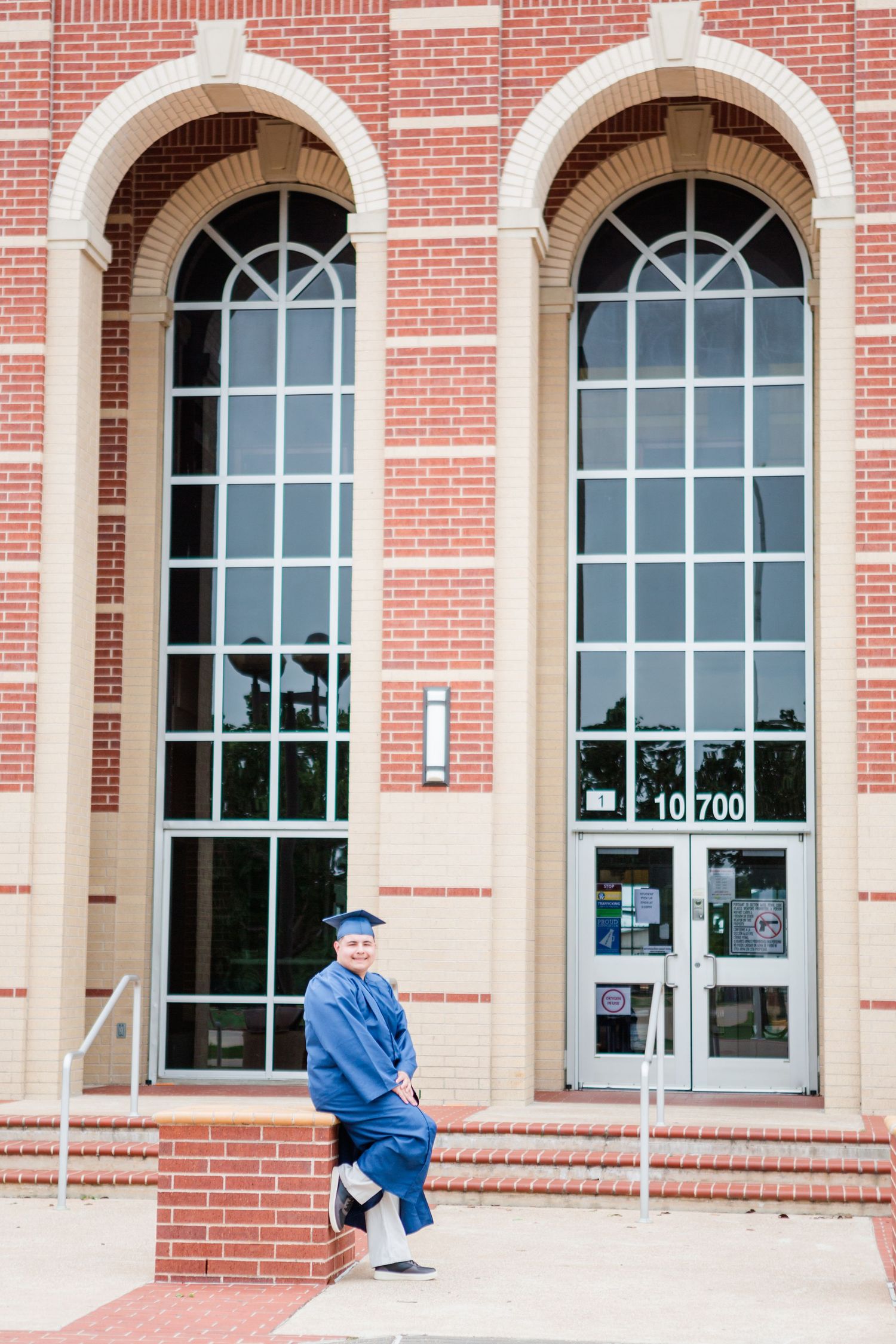 Graduate in blue cap and gown posing outside a brick building with arched windows at address 10700.