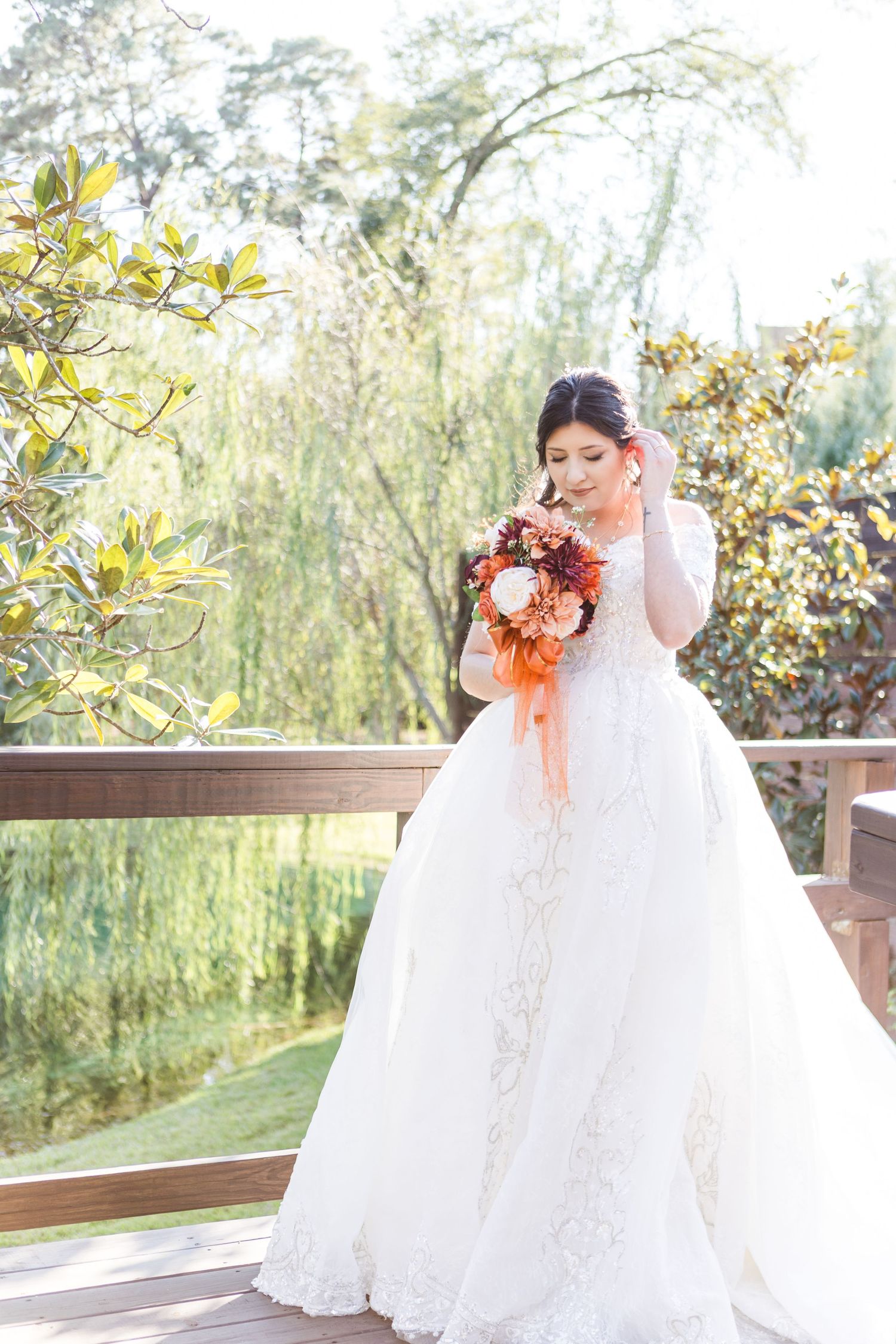 A bride in a flowing white lace gown poses outdoors on a wooden deck with willow trees in the background.