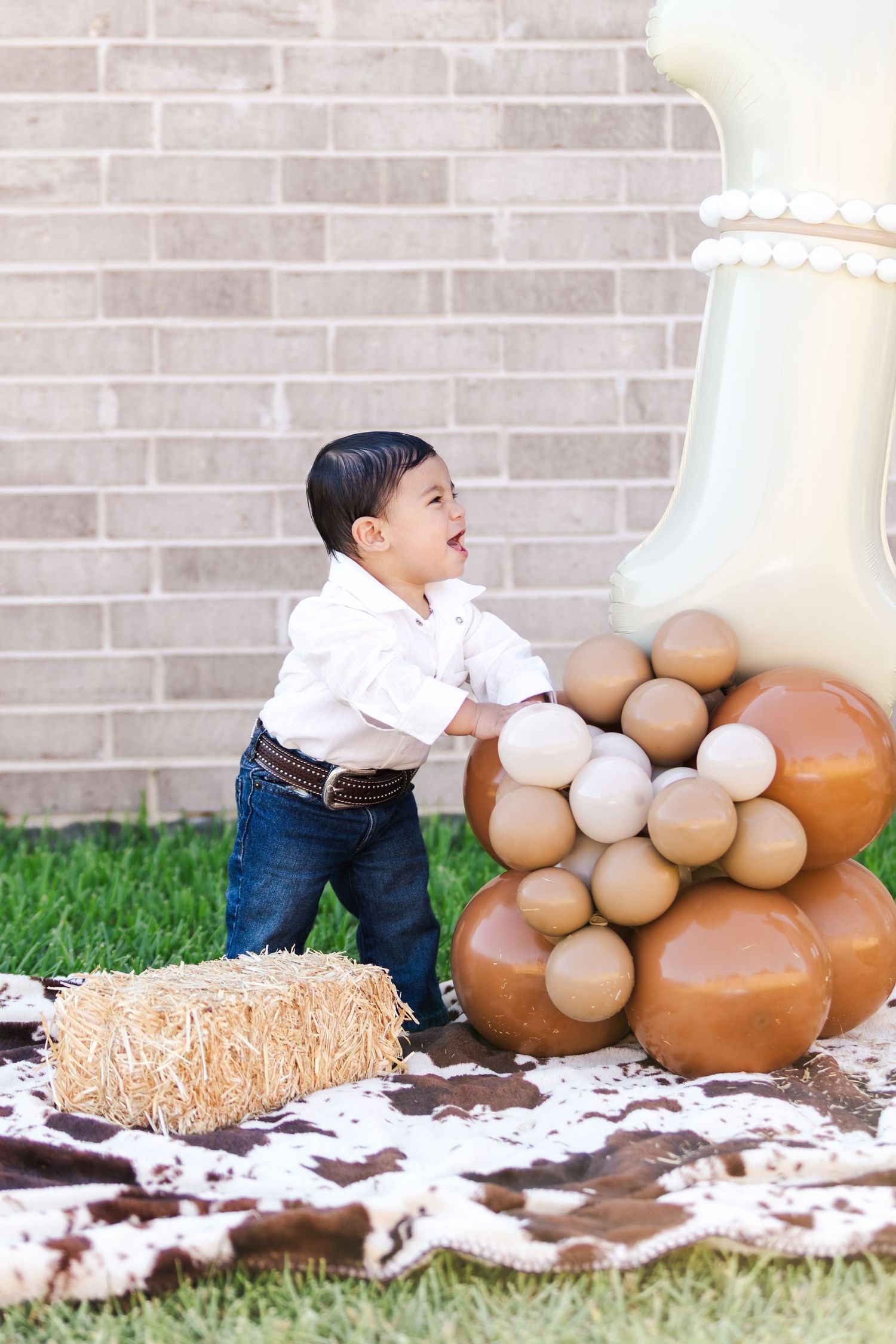 A small child in jeans and white shirt plays near autumnal decorations including balloons and hay bales.