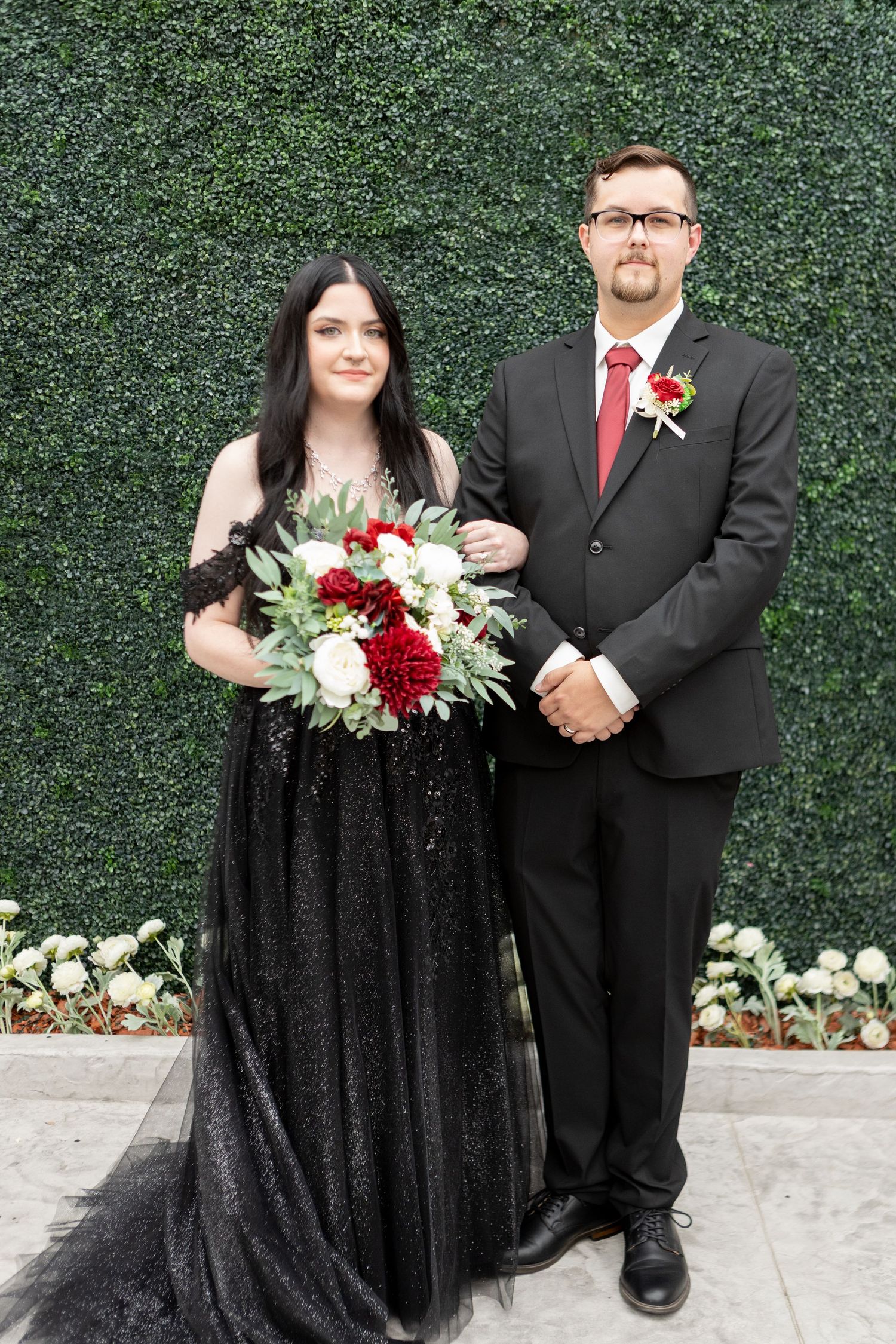 Wedding couple poses with red and white bouquet against hedge wall backdrop.