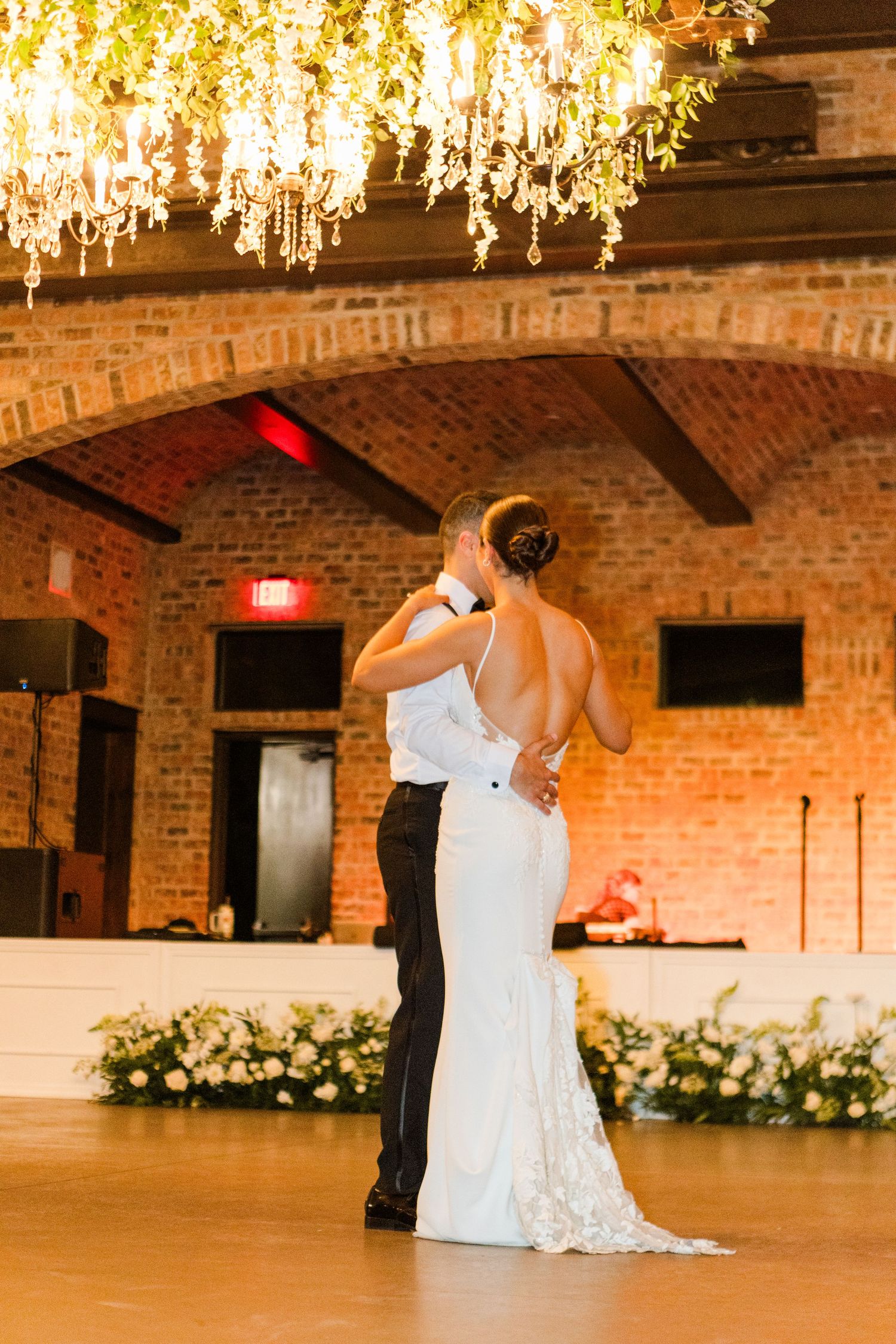 Wedding venue decorated with hanging lights and floral arrangements against brick walls.