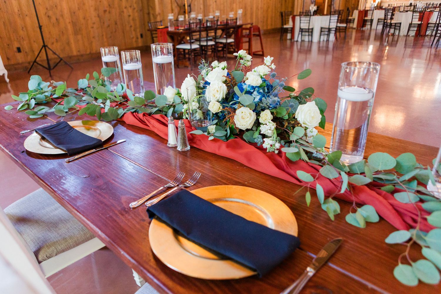Place settings at a rustic wedding reception feature gold chargers, navy napkins and eucalyptus garland on wooden tables.