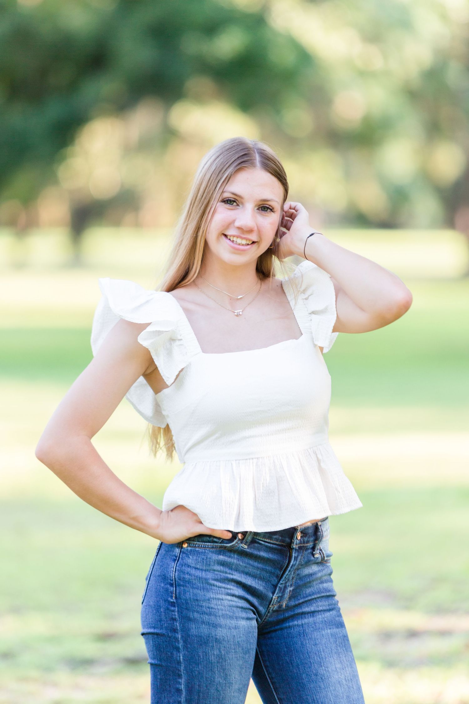 Smiling young woman in white ruffled top and jeans posing outdoors in a sunny park setting.