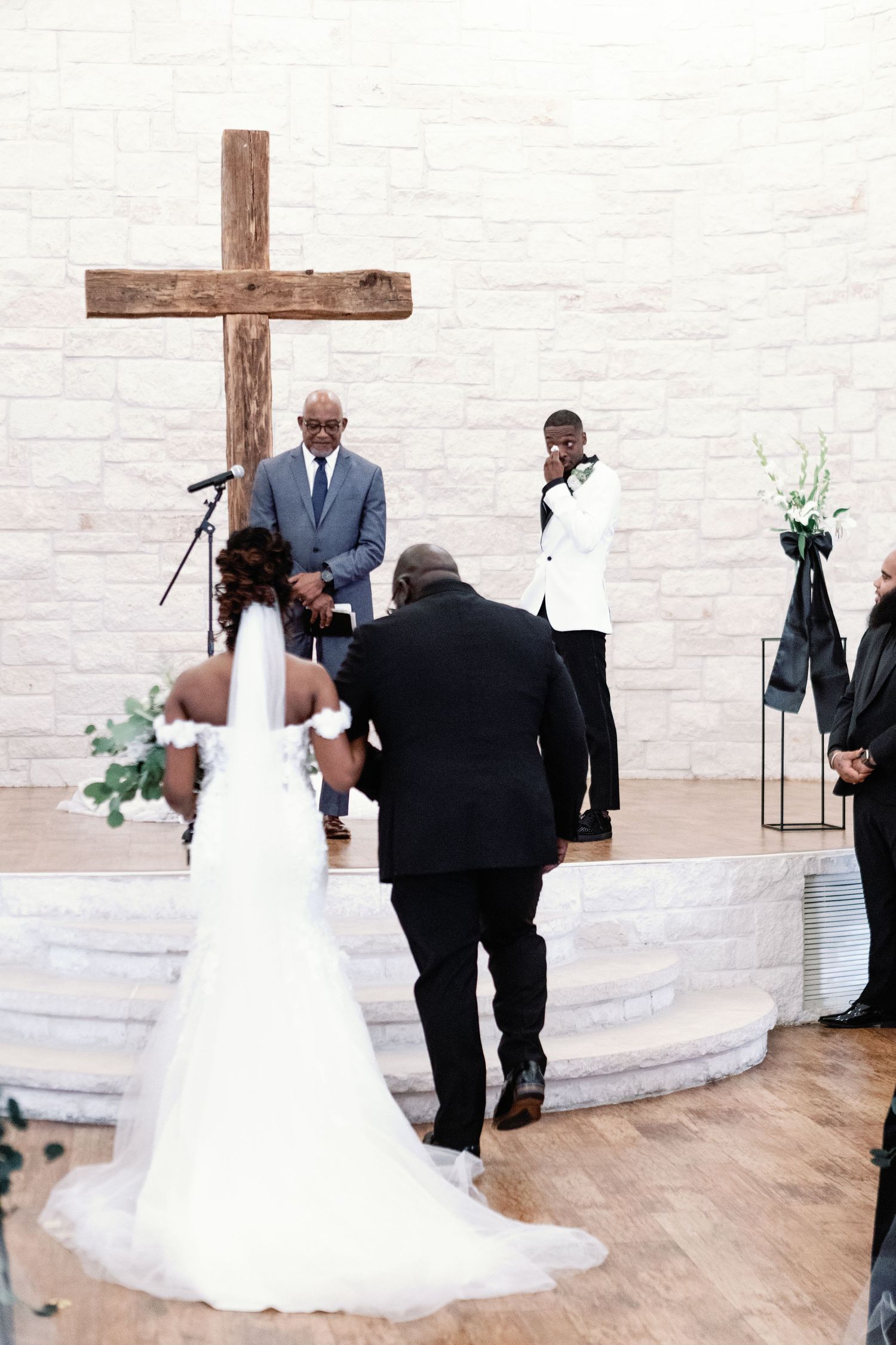 A wedding ceremony taking place in a bright white chapel with a wooden cross and flowing white wedding gown.