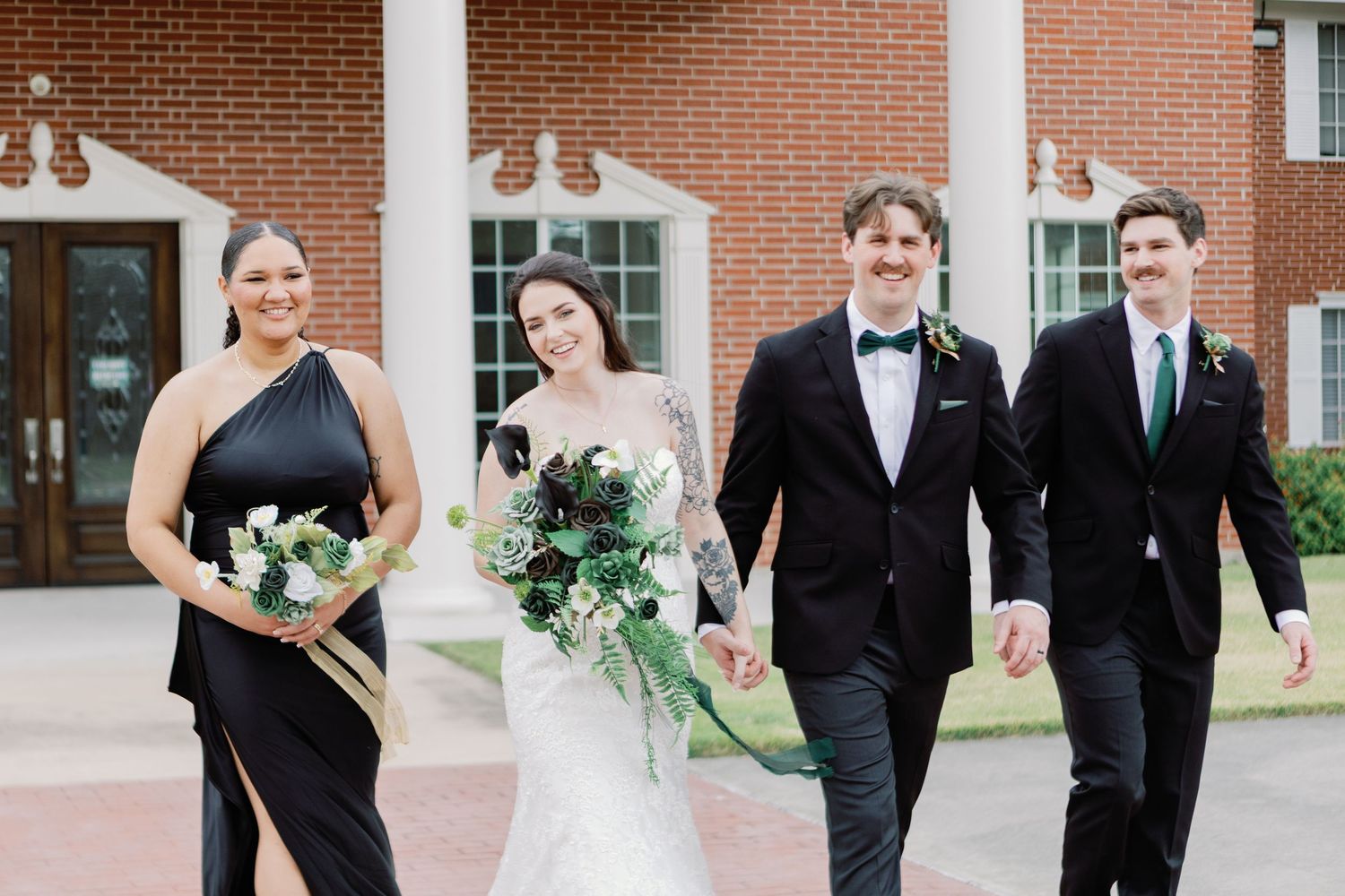 Wedding party members in formal black attire and dresses pose for portraits outside a brick building.