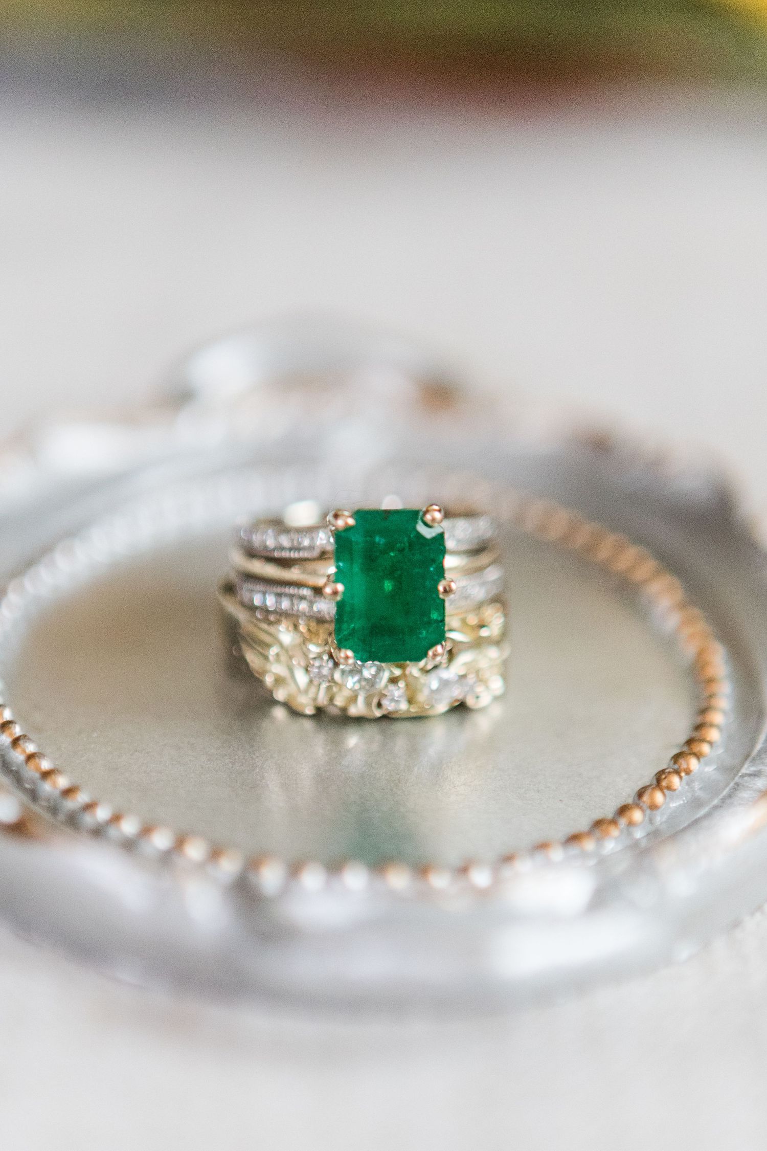 Elegant emerald and diamond engagement ring with matching pearl bracelet photographed on a light gray surface.