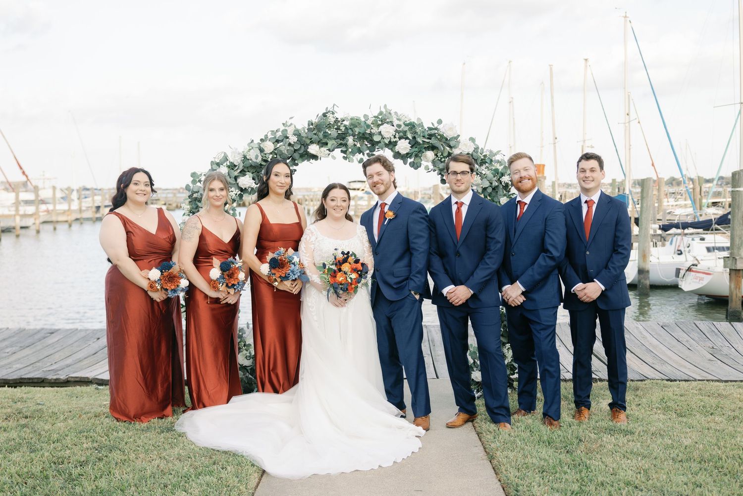 A wedding party in navy blue suits and rust colored dresses poses together outdoors by a waterfront venue.