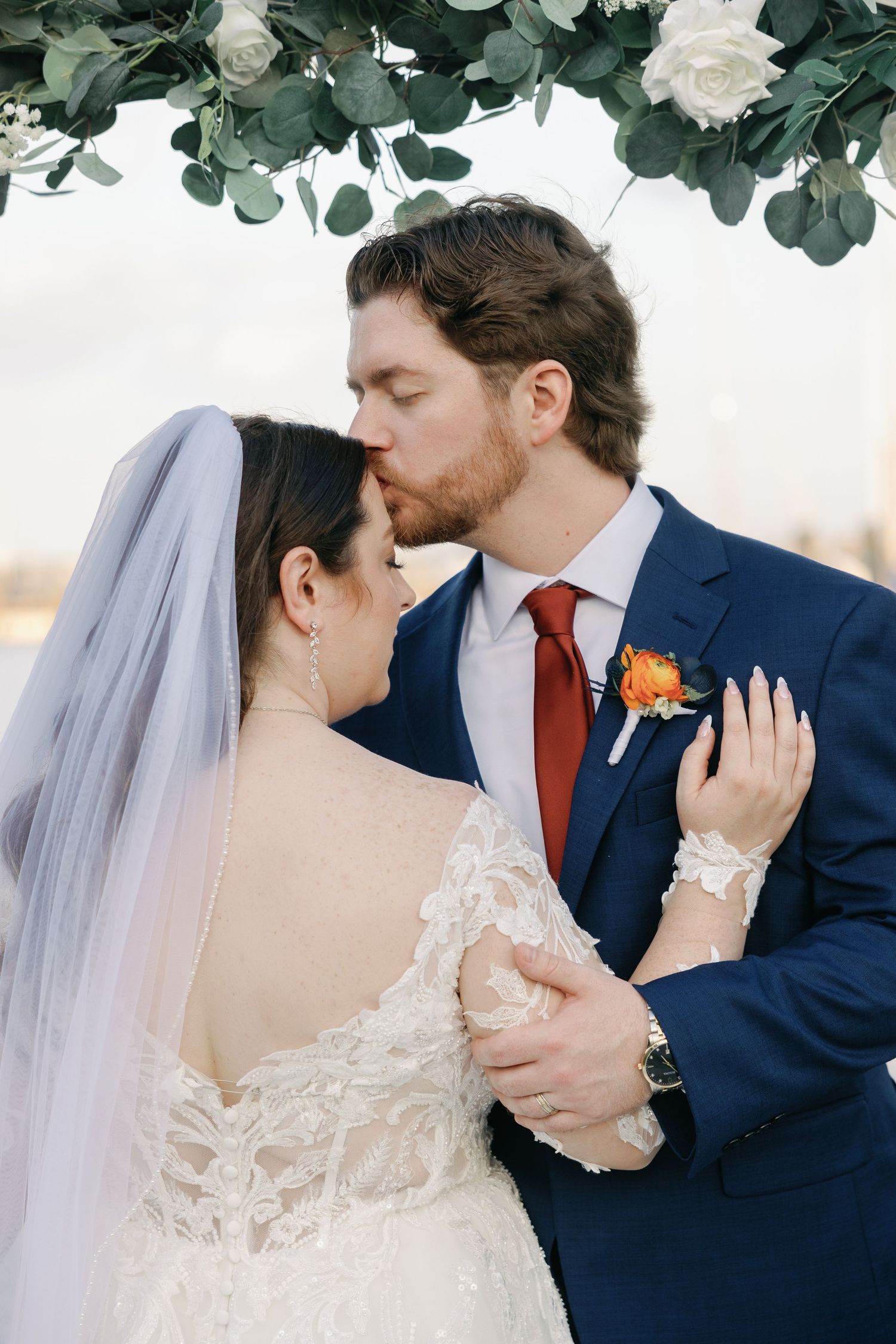Romantic wedding moment under eucalyptus arch with bride in lace gown and groom in navy suit with orange boutonniere.