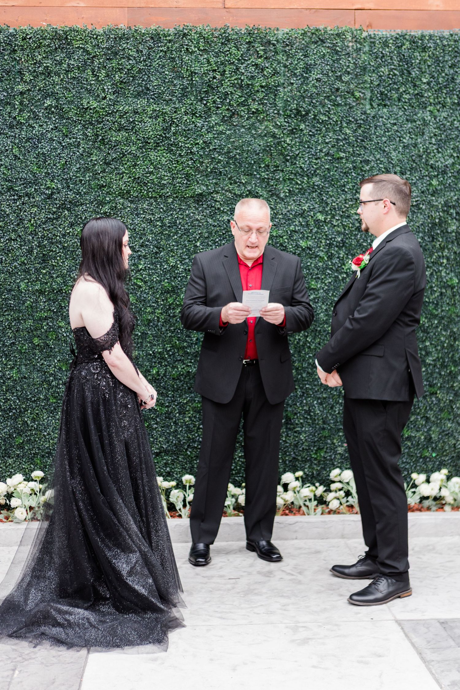 Wedding party in black attire gathered for photos against decorative green hedge wall.