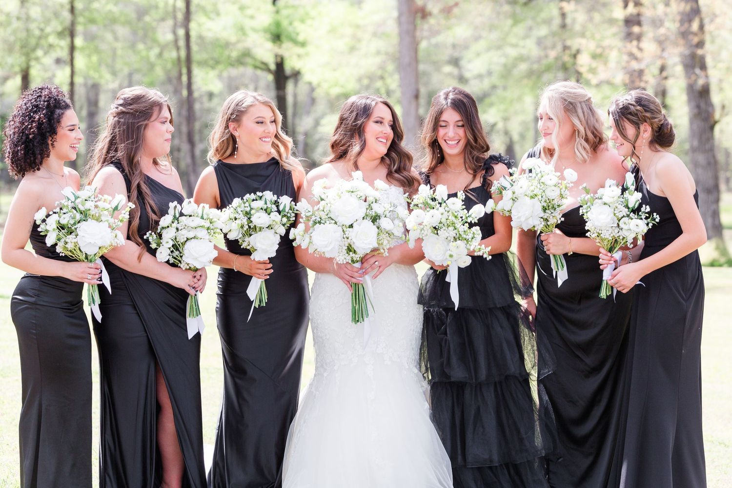 Bridesmaids in black dresses holding white floral bouquets pose together outdoors.