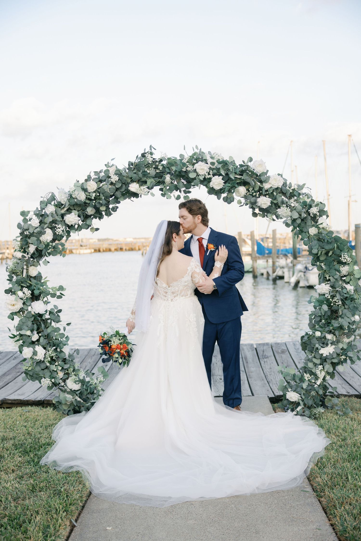 A romantic waterfront wedding ceremony under a lush floral arch with white and green decorations.