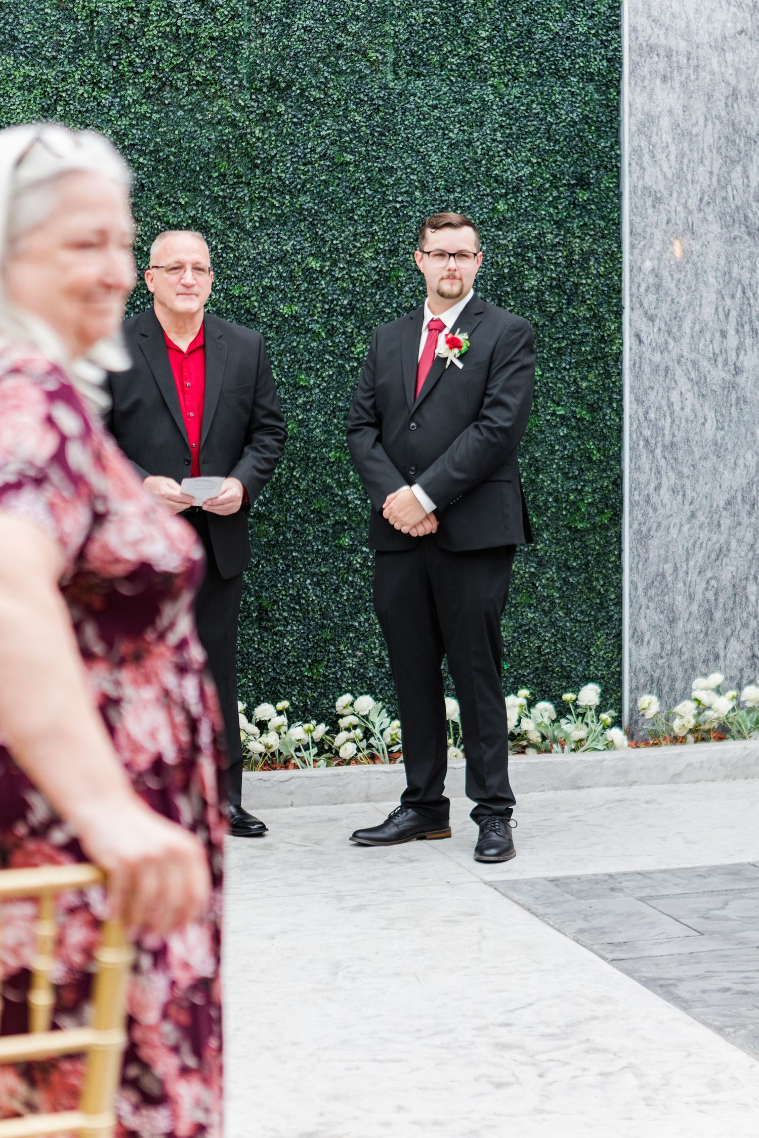 Wedding party members in formal attire pose against green hedge wall.