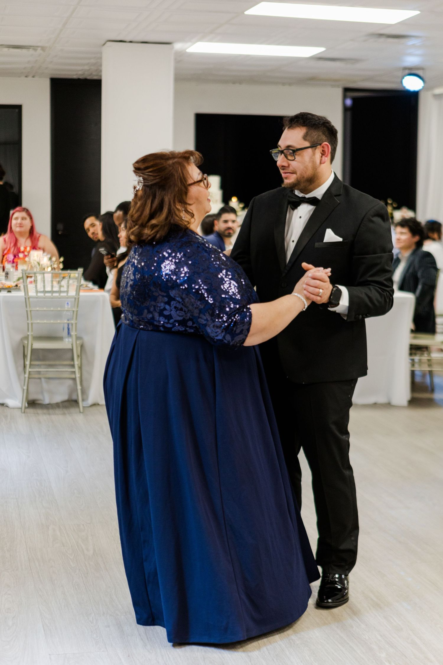 A couple in navy blue and black formal attire dance together at a wedding reception.