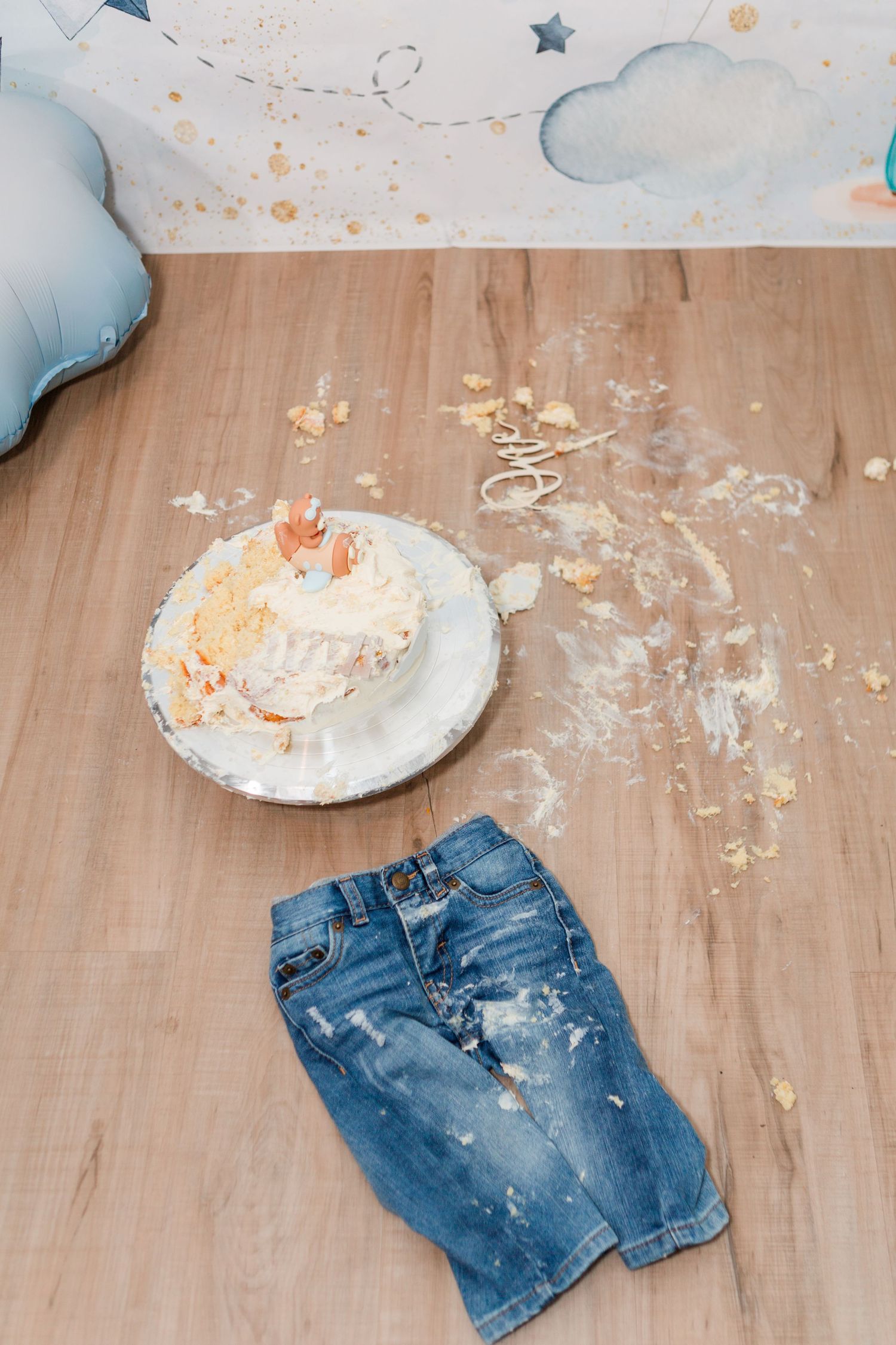 Demolished birthday cake on a plate next to denim shorts on wooden floor.