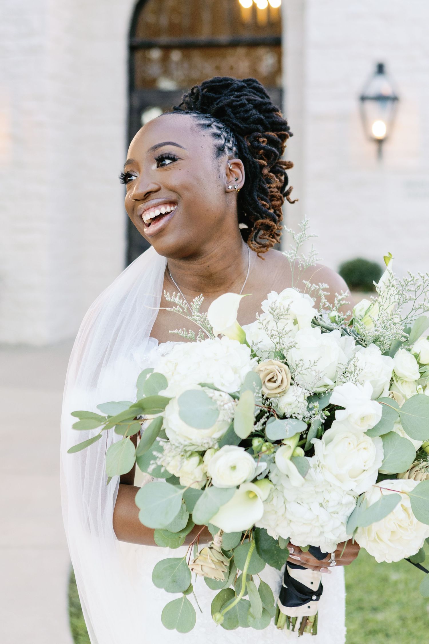 A bride in a white wedding dress holds a beautiful bouquet of white roses and eucalyptus leaves while laughing joyfully.