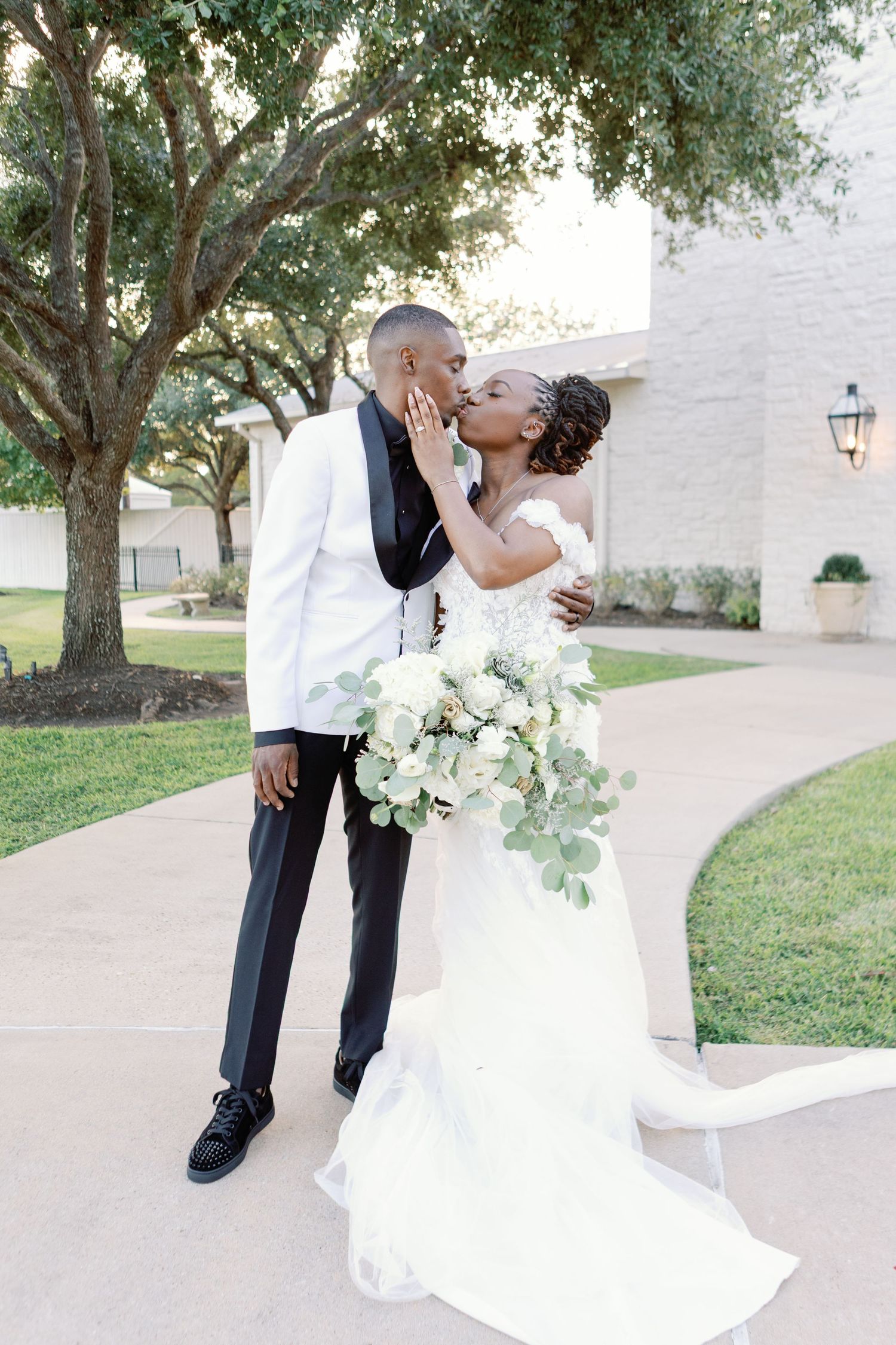 Romantic wedding couple embracing outdoors near white building and trees.