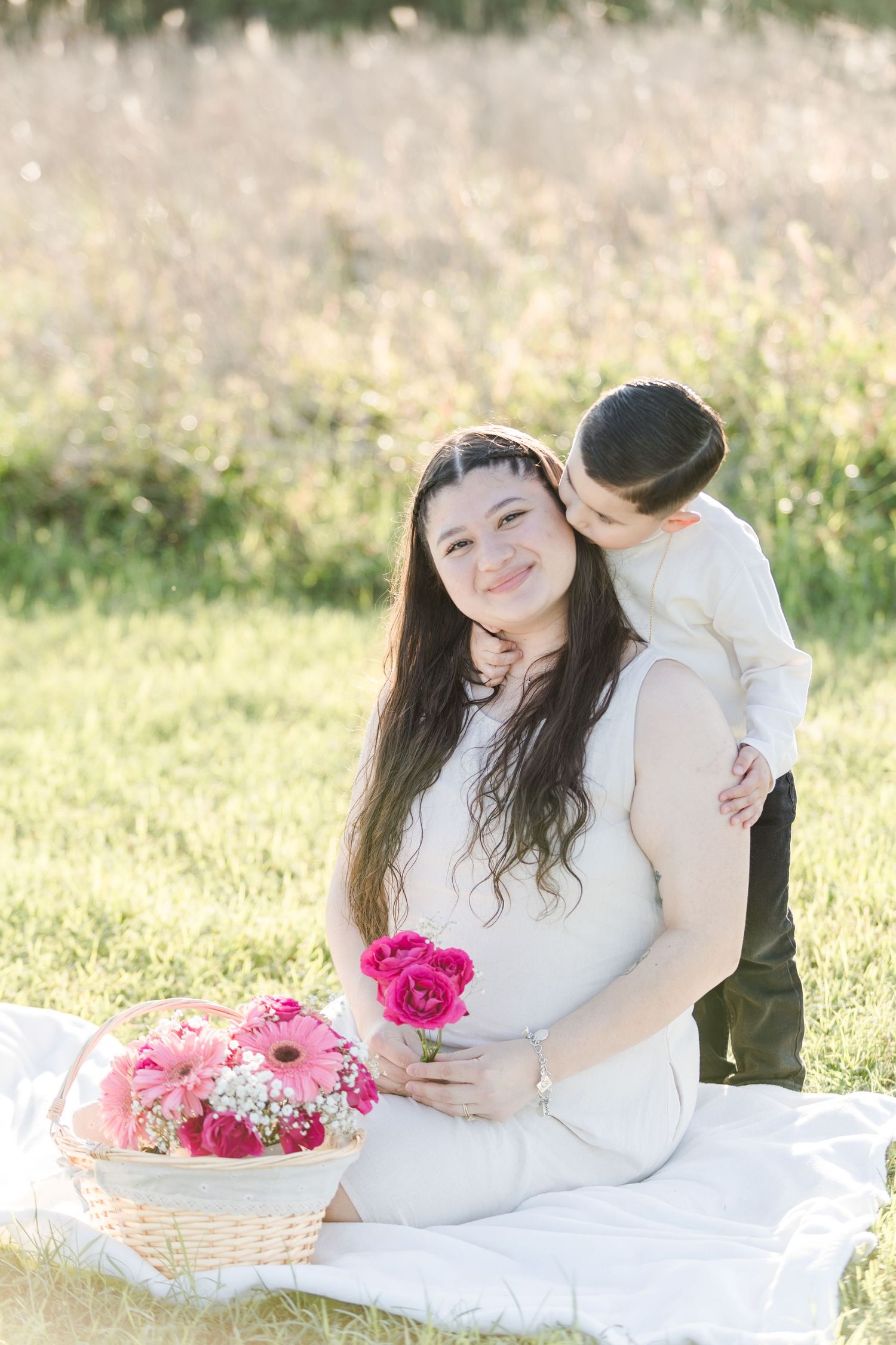 Couple in white poses in a sunny field with pink flowers for romantic maternity photos.