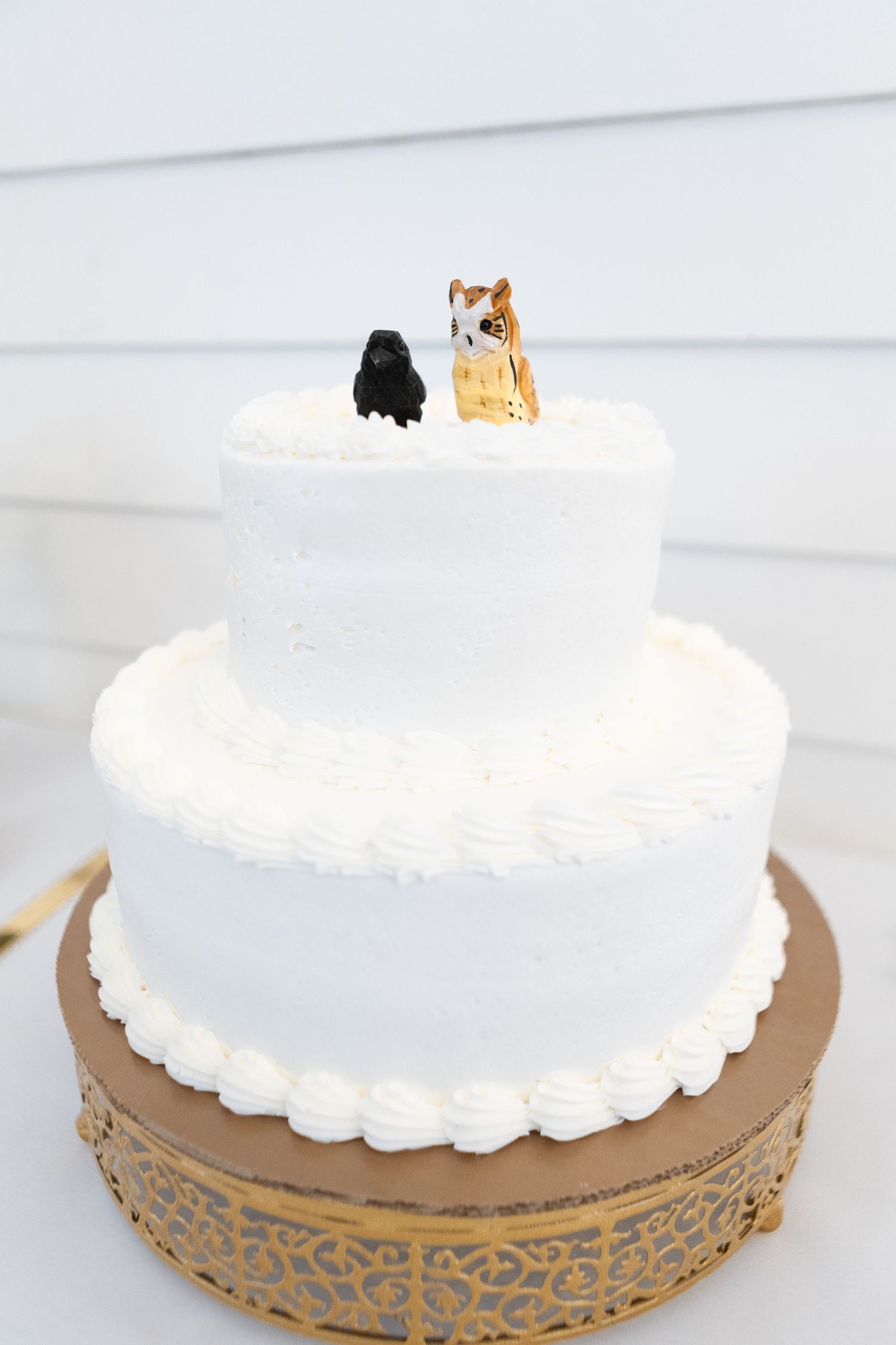 White frosted wedding cake with cat toppers displayed on an ornate gold stand.