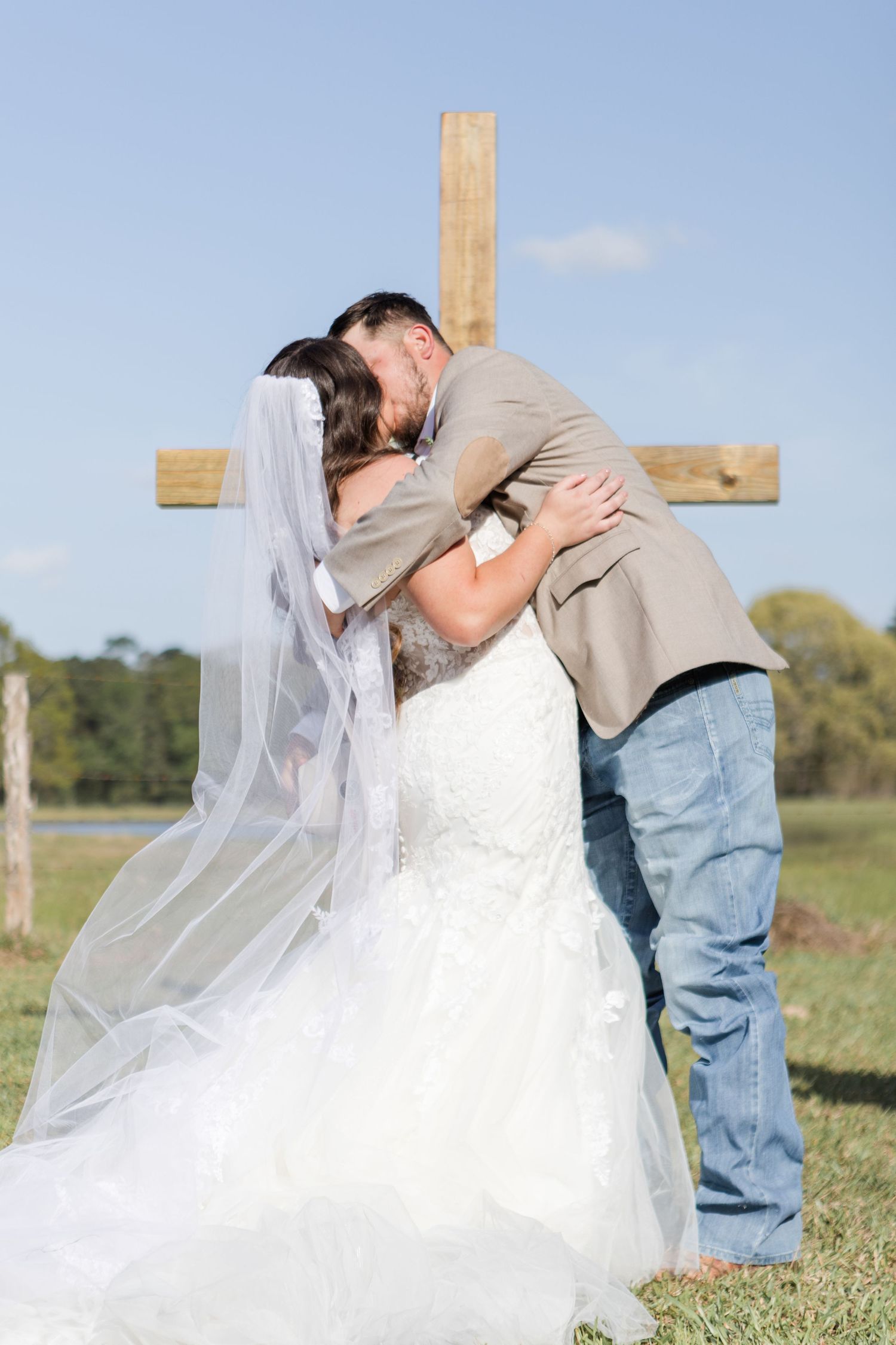 Wedding couple embracing near a wooden cross in an outdoor field setting.