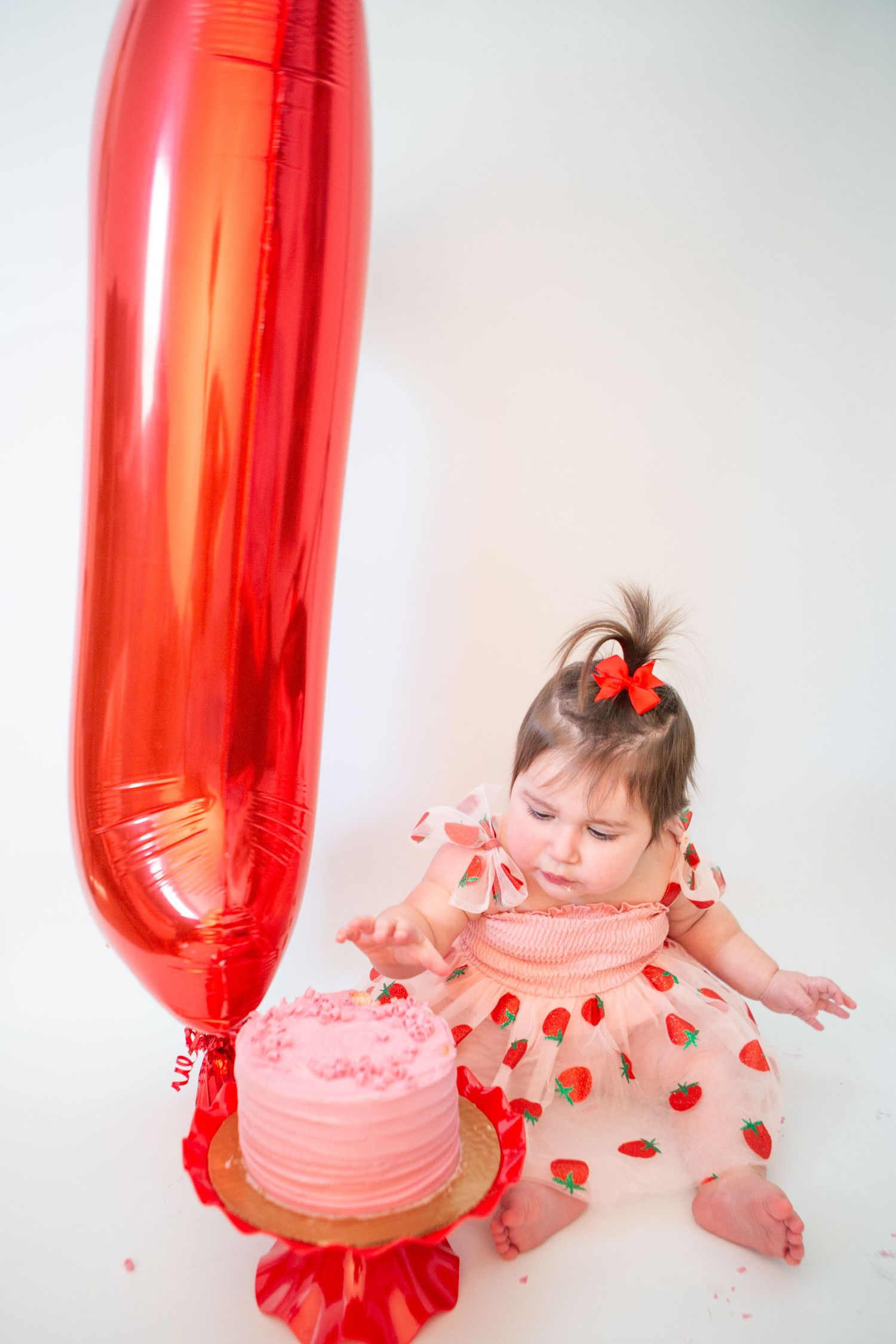Baby in heart-print outfit reaching for pink cake with red balloon decoration.