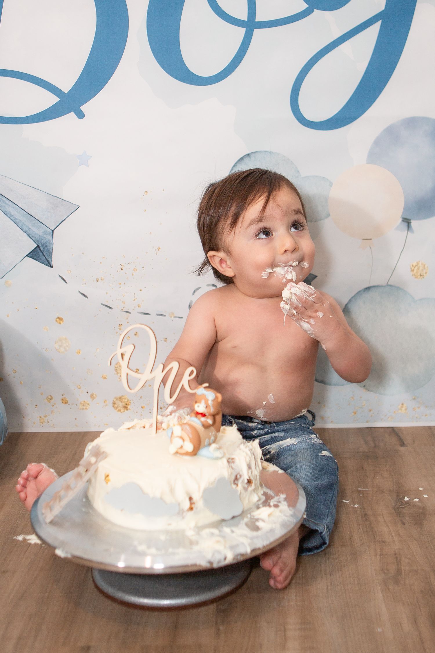 Messy cake smash scene with baby and white frosted cake on wooden floor.