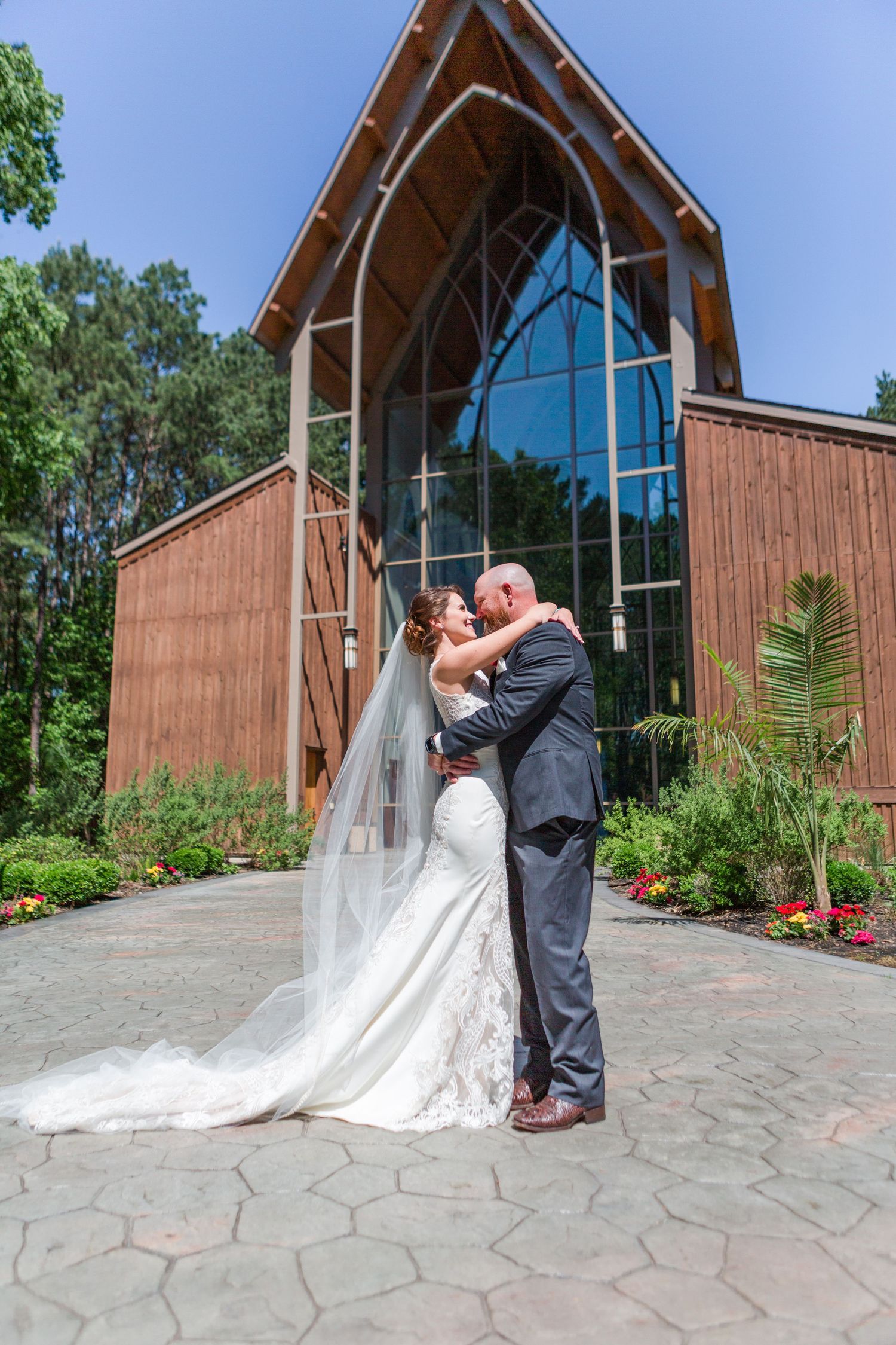 A couple shares a romantic kiss outside a rustic wooden chapel with large windows and stone pavement.