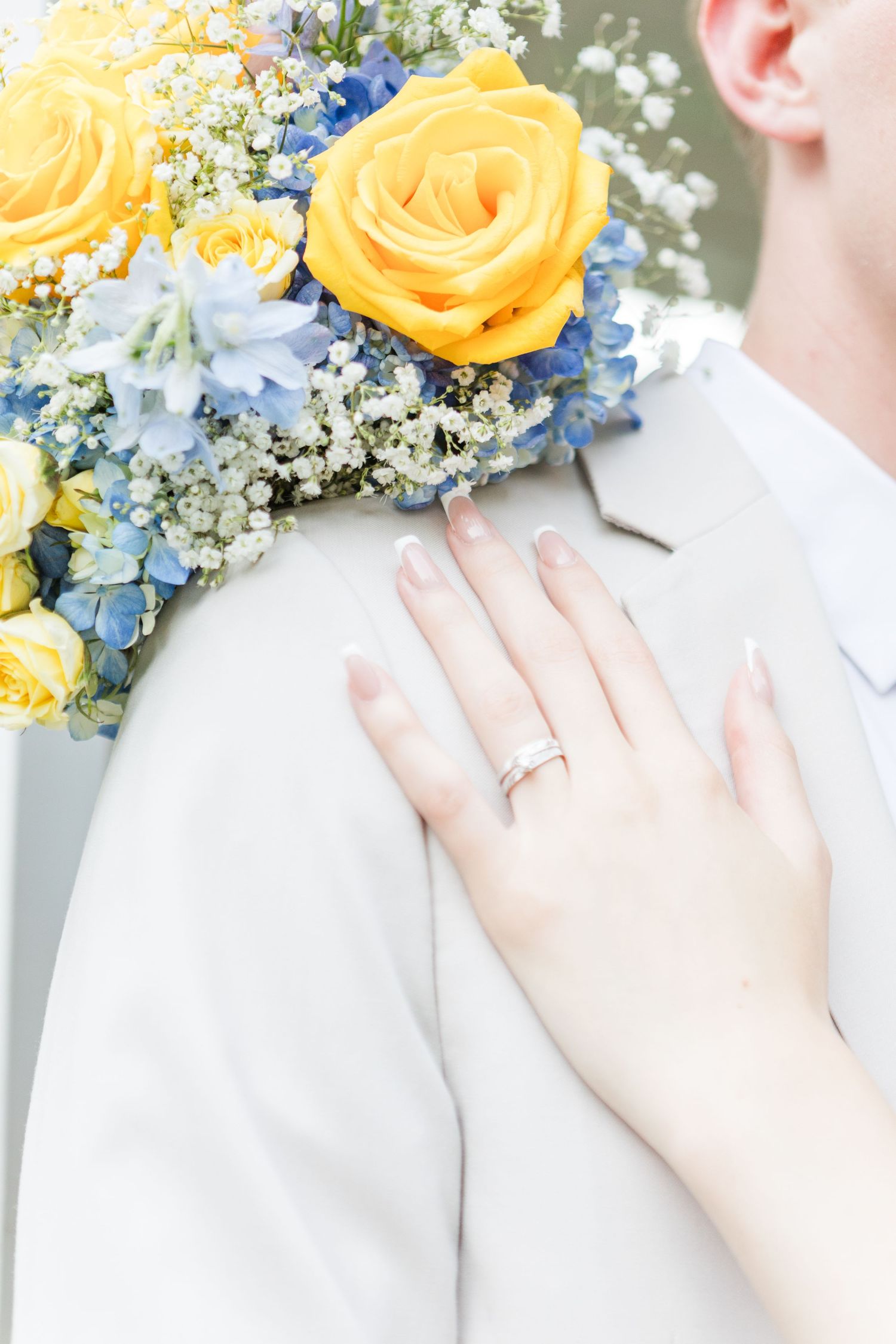 Yellow and blue floral bouquet with hand displaying engagement ring on gray suit.