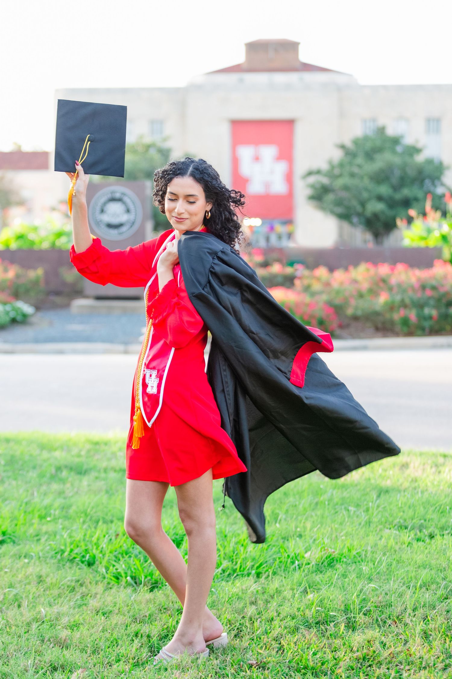 A young woman in red dress and black graduation gown holds cap aloft on grass near University of Houston building.