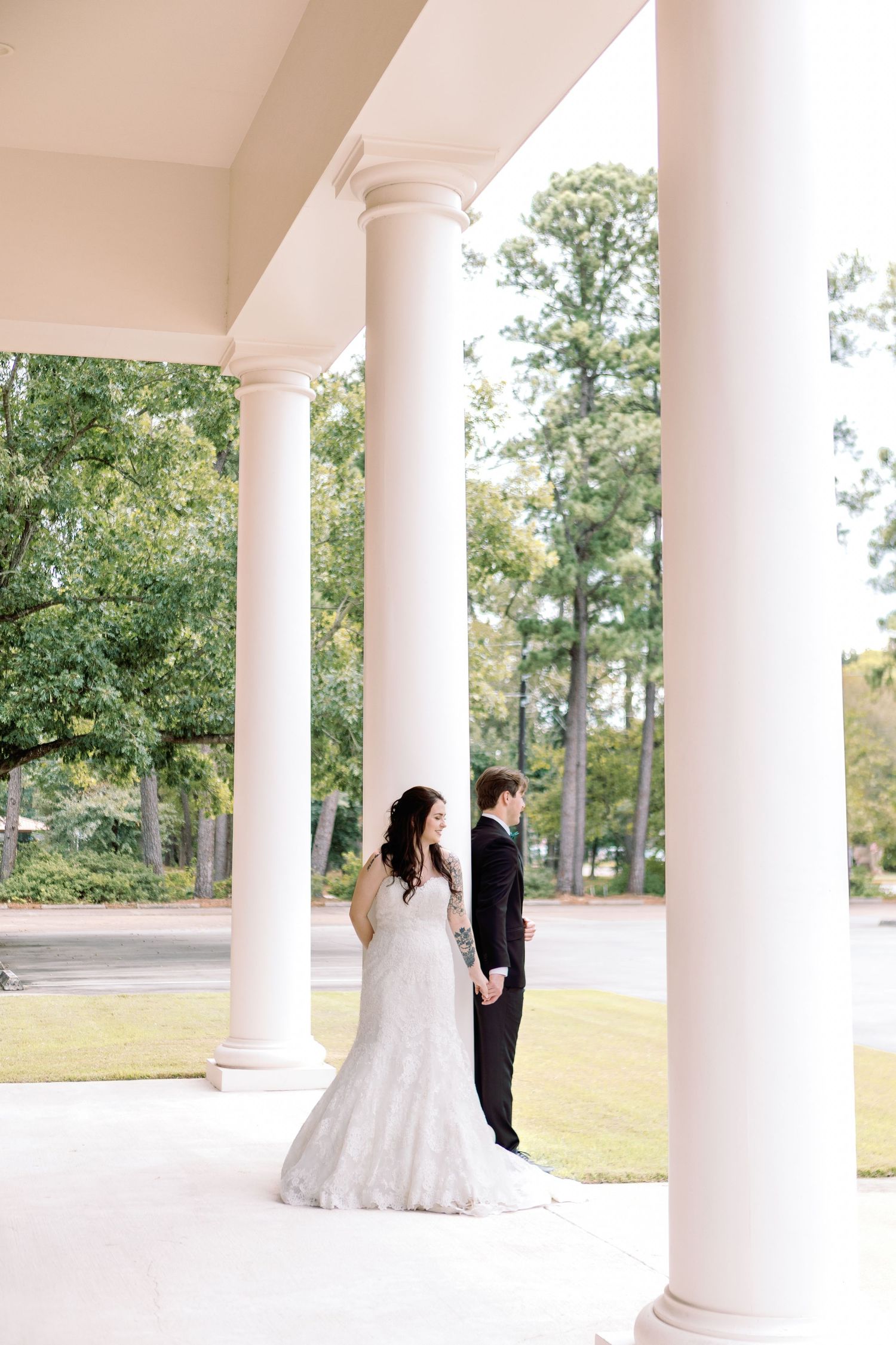 A couple shares a romantic moment between elegant white columns on a porch overlooking a scenic garden landscape.