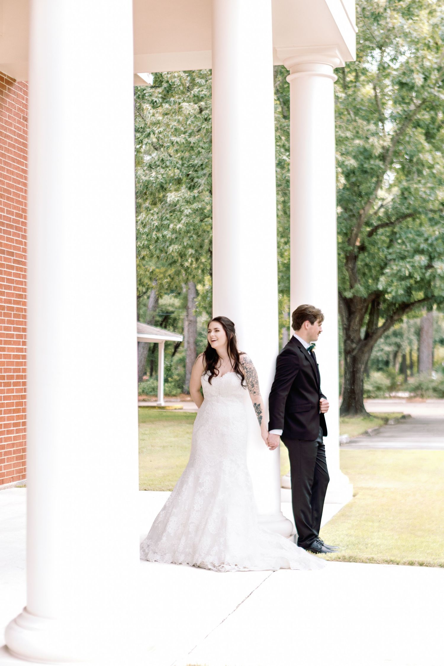 A bride and groom stand between white columns on a porch, creating a romantic wedding portrait.