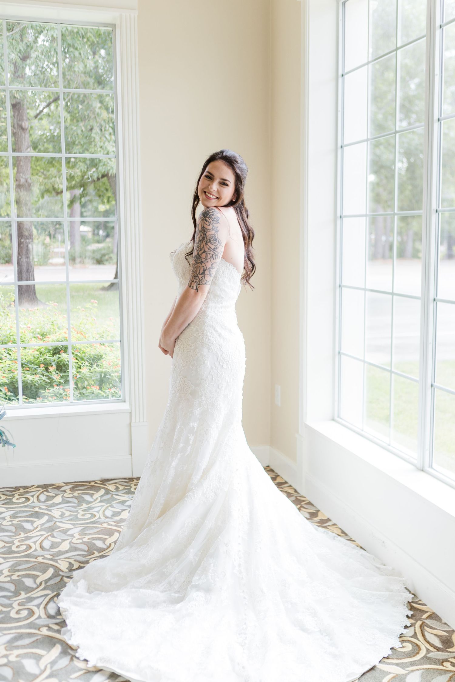 Bride in lace wedding gown with long train poses by sunlit windows on patterned floor.
