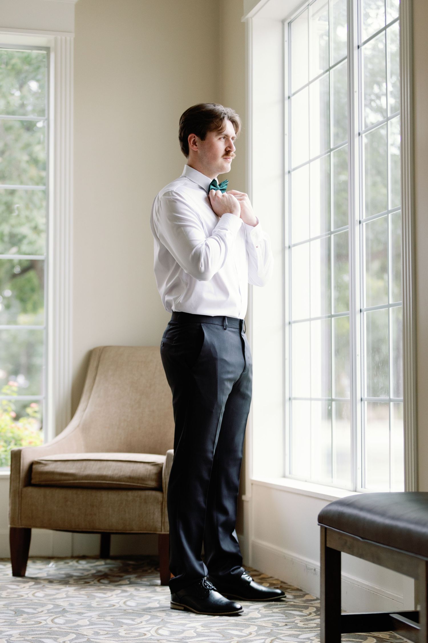 Man in formal attire stands pensively by bright windows in an elegant room with beige furniture.
